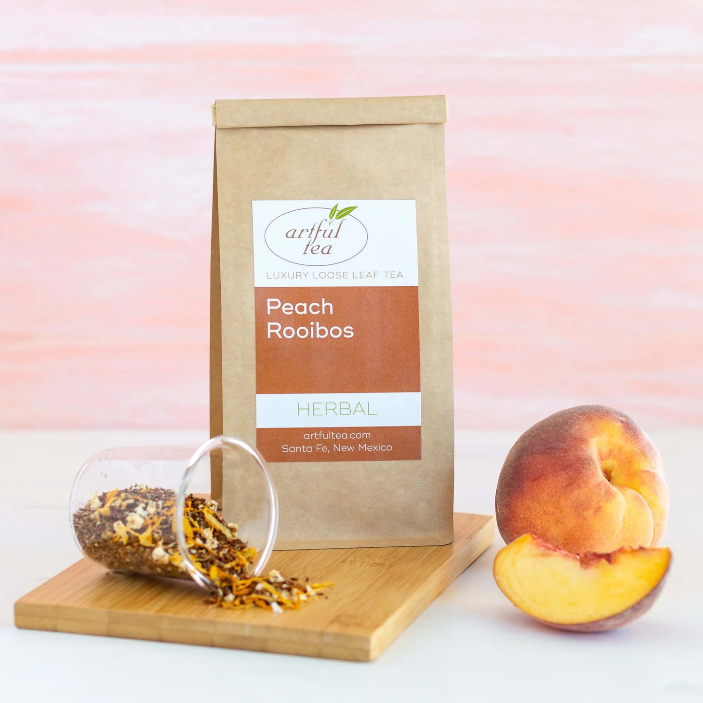 Peach Rooibos Herbal Tea shown packaged in a brown Kraft bag next to a clear cup spilling loose leaf tea on top of a wooden square with peaches next to it.