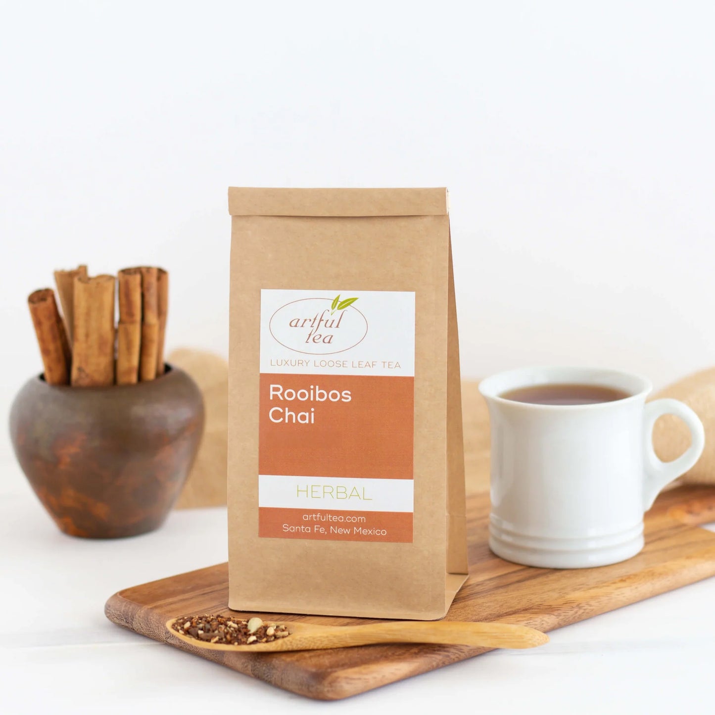 Rooibos Chai Herbal Tea shown packaged in a brown Kraft bag next to a white mug full of brewed tea and a spoon of loose leaf tea on a wooden platter.