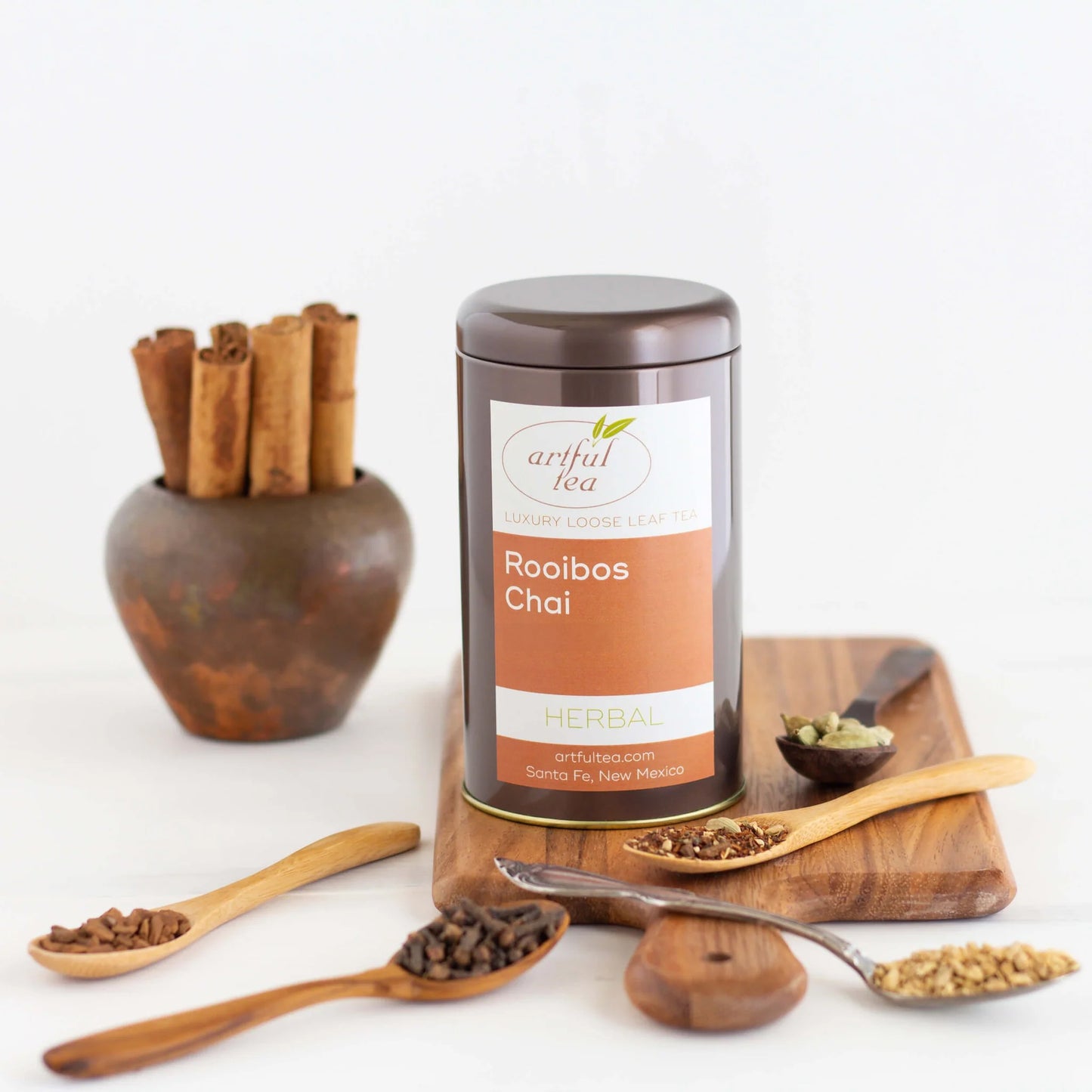 Rooibos Chai Herbal Tea shown packaged in a brown tin on a wooden board with ingredients in different spoons nearby.