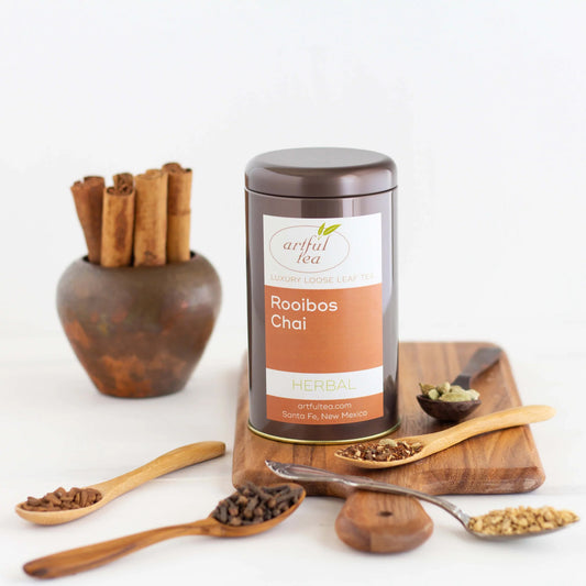 Rooibos Chai Herbal Tea shown packaged in a brown tin on a wooden board with ingredients in different spoons nearby.