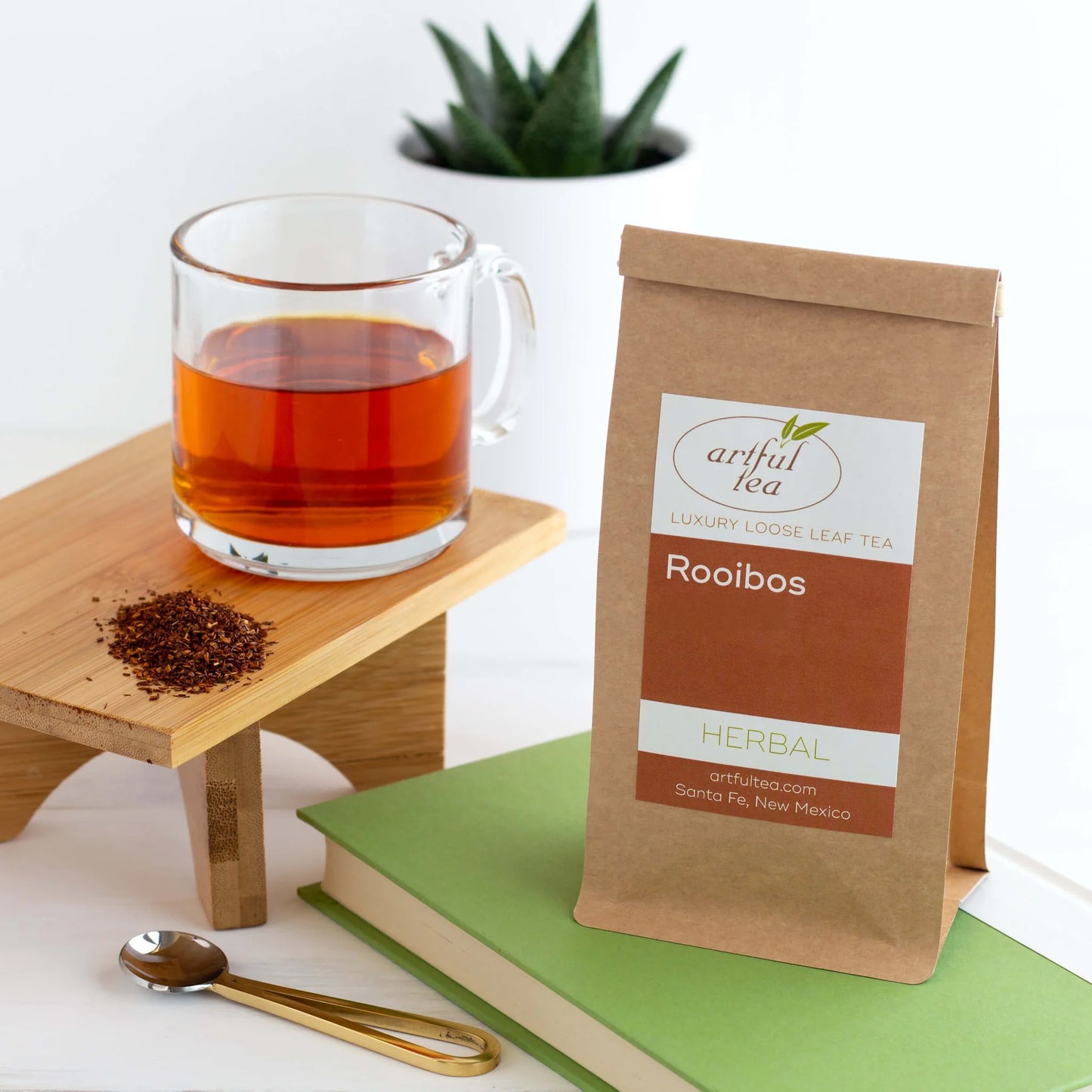 Rooibos Herbal Tea shown packaged in a brown Kraft bag on top of a green book. Beside it is a clear mug full of brewed tea on top of a wooden platter.