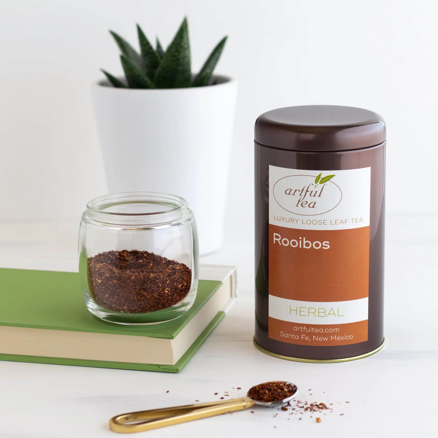 Rooibos Herbal Tea shown packaged in a brown tin next to a clear jar of loose leaf tea on top of a green book. In front is a scoop of loose tea and in the back is a plant with a white pot.