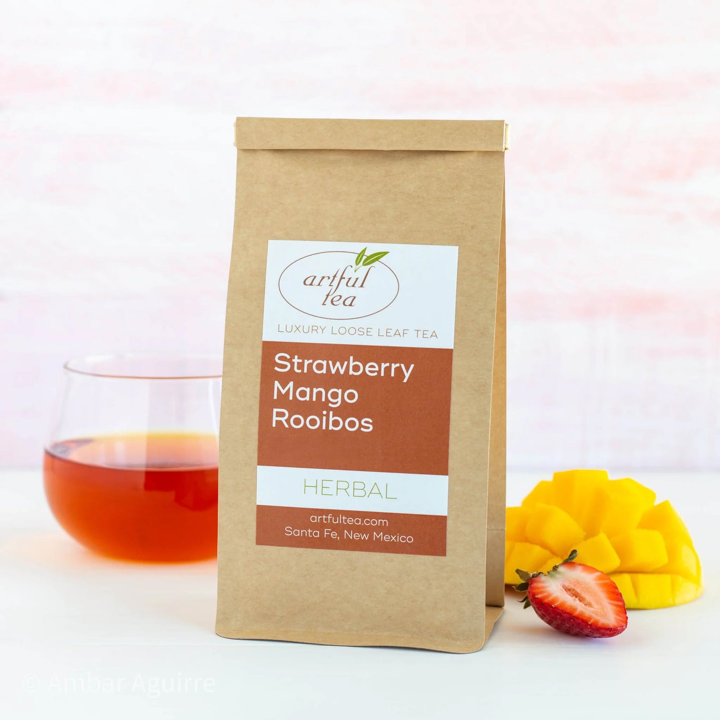 Strawberry Mango Rooibos Herbal Tea shown packaged in a brown Kraft bag next to a clear cup of brewed tea and ingredients.