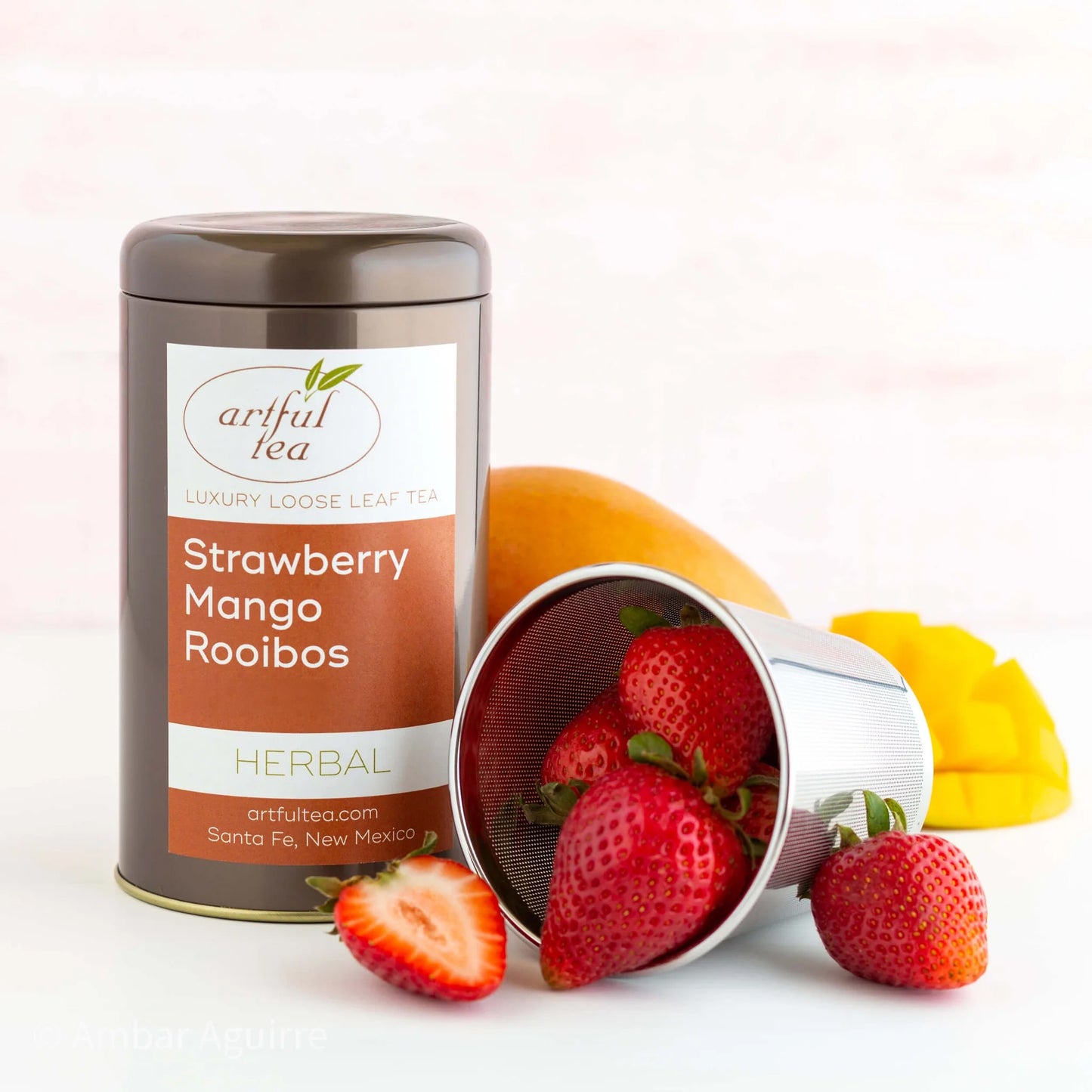Strawberry Mango Rooibos Herbal Tea shown packaged in a brown tin next to a strainer full of strawberries with mangos behind it.