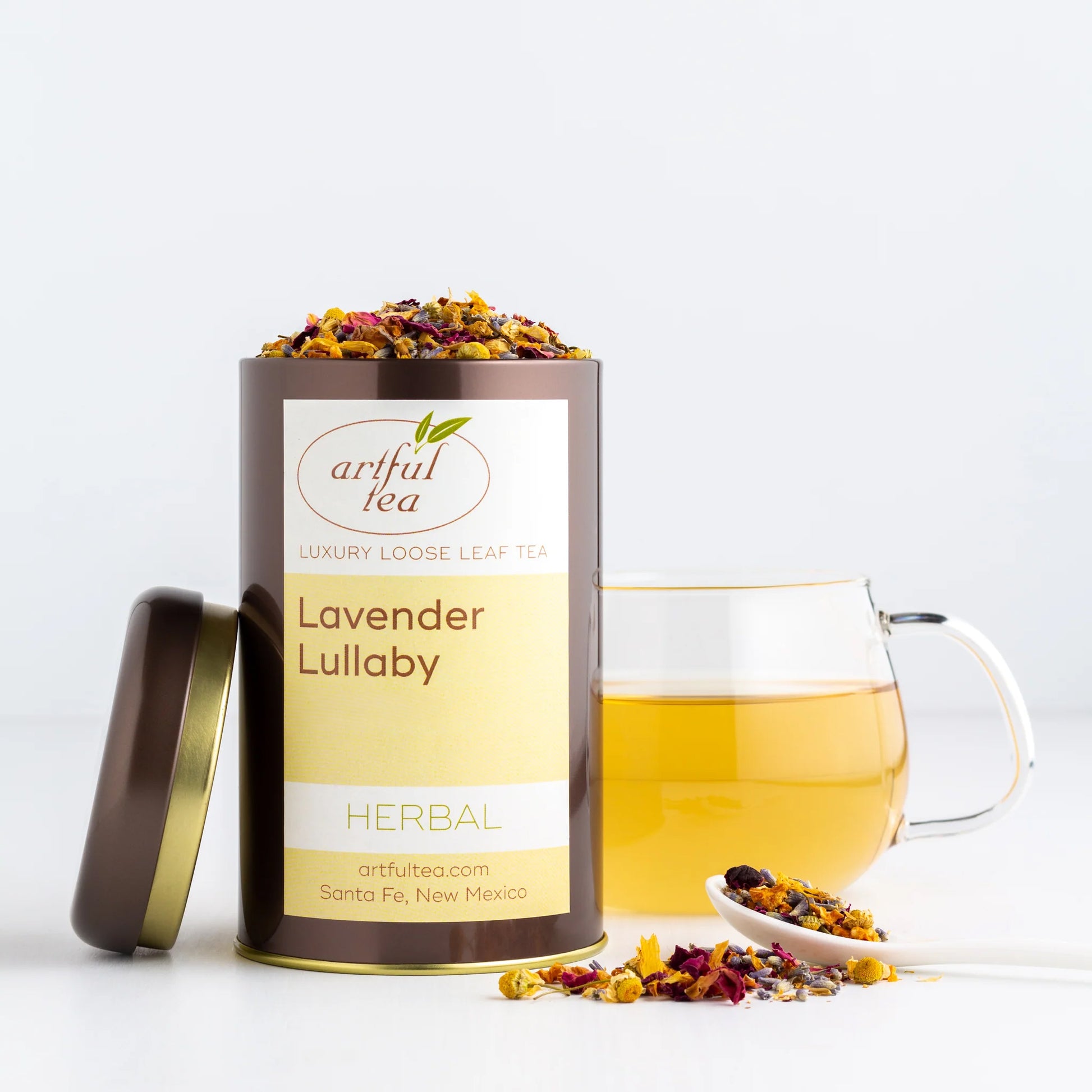 Lavender Lullaby Herbal Tea shown packaged in a brown tin with the lid off next to a clear mug of brewed tea and a white spoon of loose leaf tea.