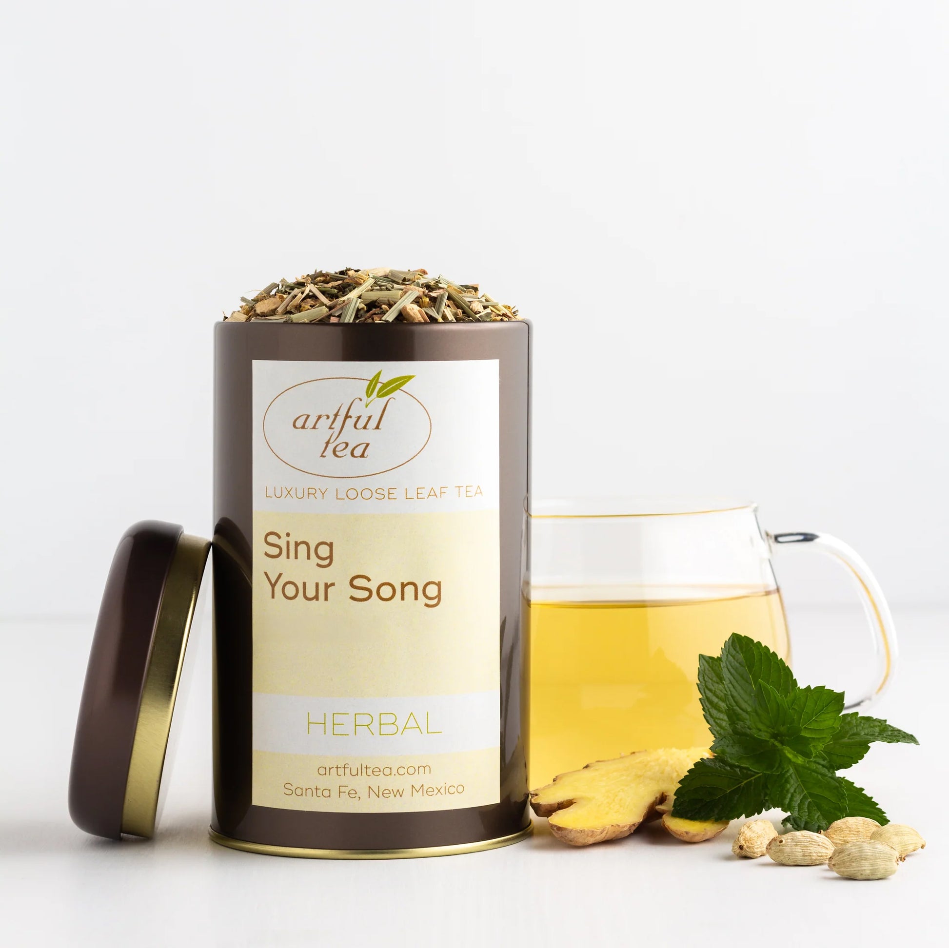 Sing Your Song Herbal Tea shown packaged in a brown tin with the lid off next to a clear mug of brewed tea and ingredients.
