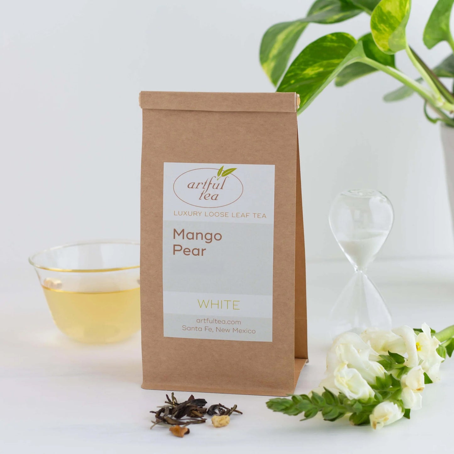 Mango Pear White Tea shown packaged in a brown Kraft bag. Next to it is a clear hourglass, white flowers, and a clear cup full of brewed tea.