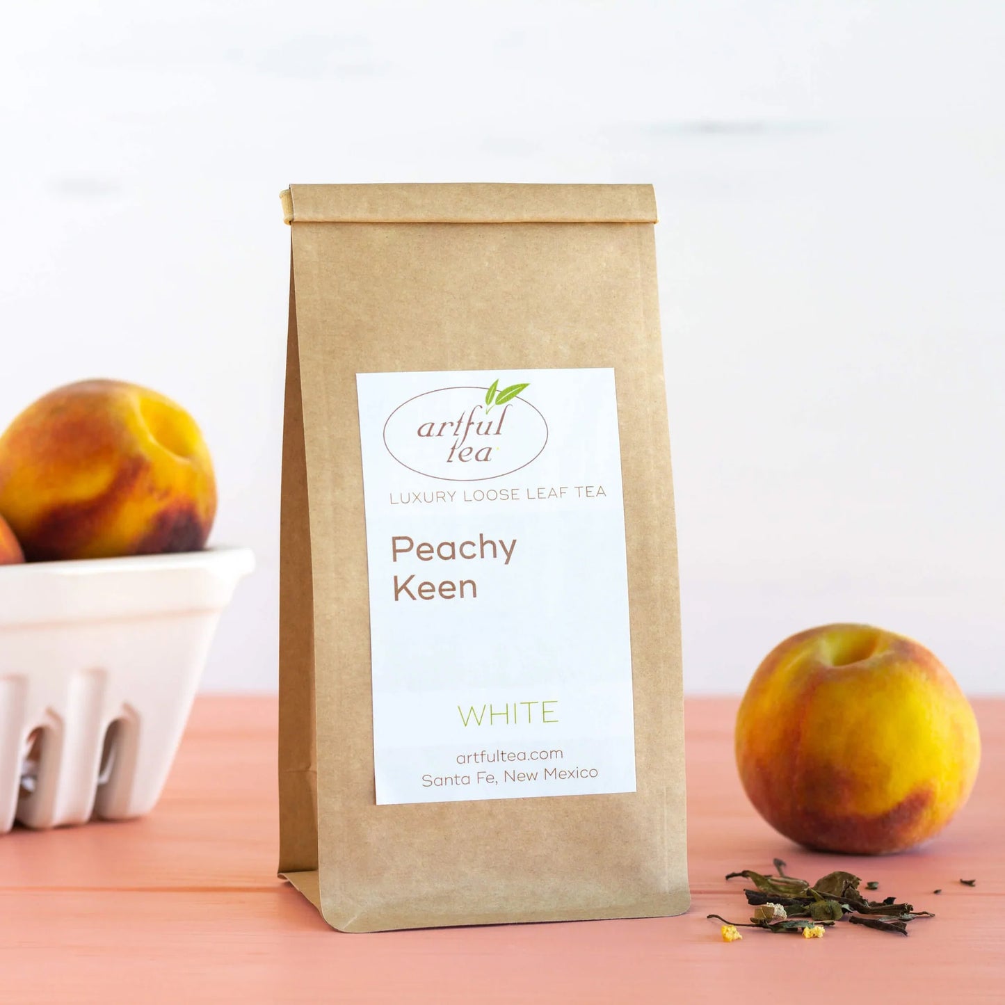 Peachy Keen White Tea shown packaged in a brown Kraft bag with peaches nearby and a small pile of loose leaf tea.