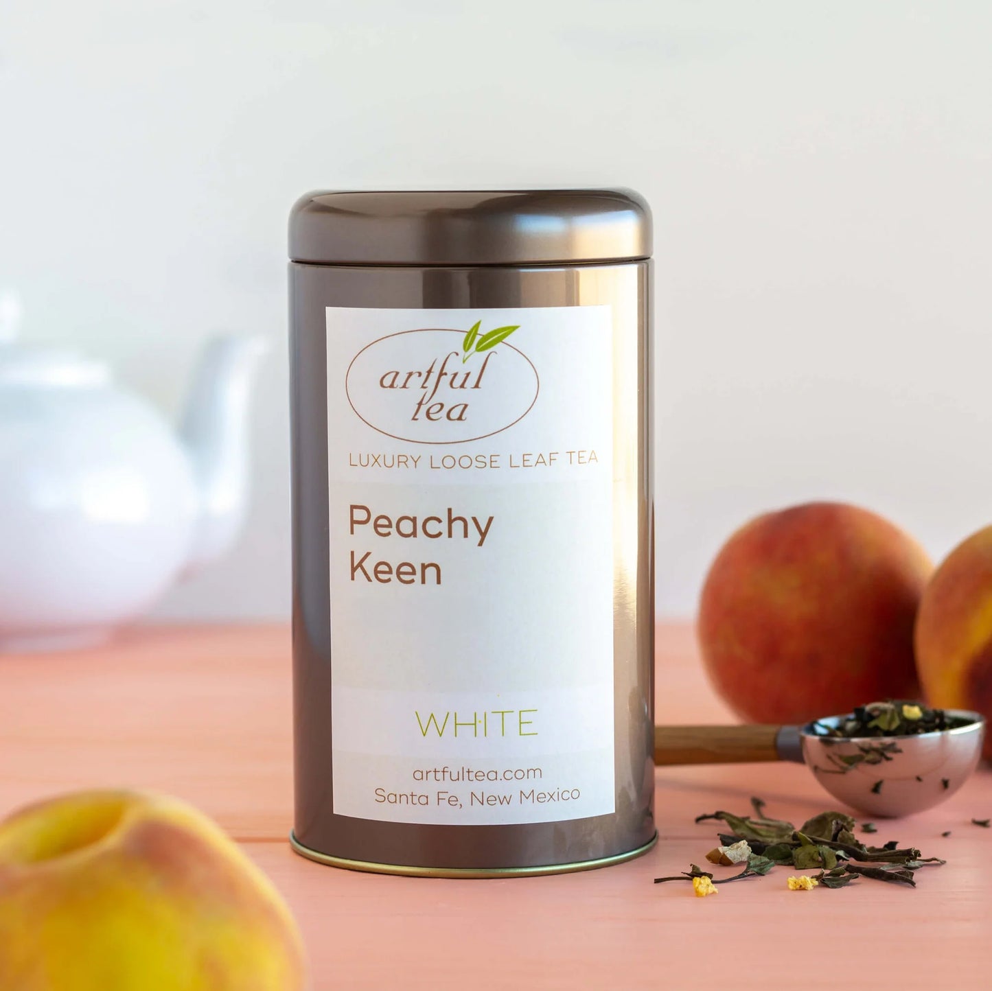 Peachy Keen White Tea shown packaged in a brown tin with a scoop of l rose leaf tea next to it and peaches around it.