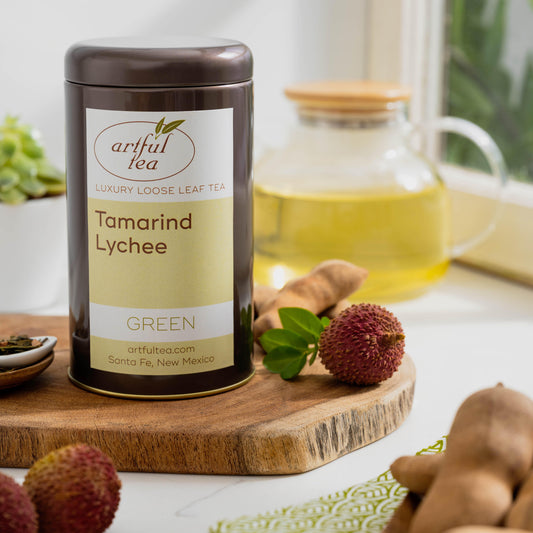 Tamarind Lychee Green Tea shown packaged in a brown tin on top of a wooden plank with ingredients nearby. In the background is a clear teapot full of brewed tea.
