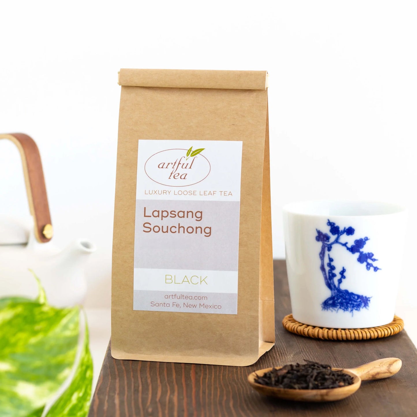Lapsang Souchong Black Tea shown packaged in a brown Kraft bag. Beside it is a white and blue bonsai cup on a woven coaster and wooden scoop full of loose leaf tea