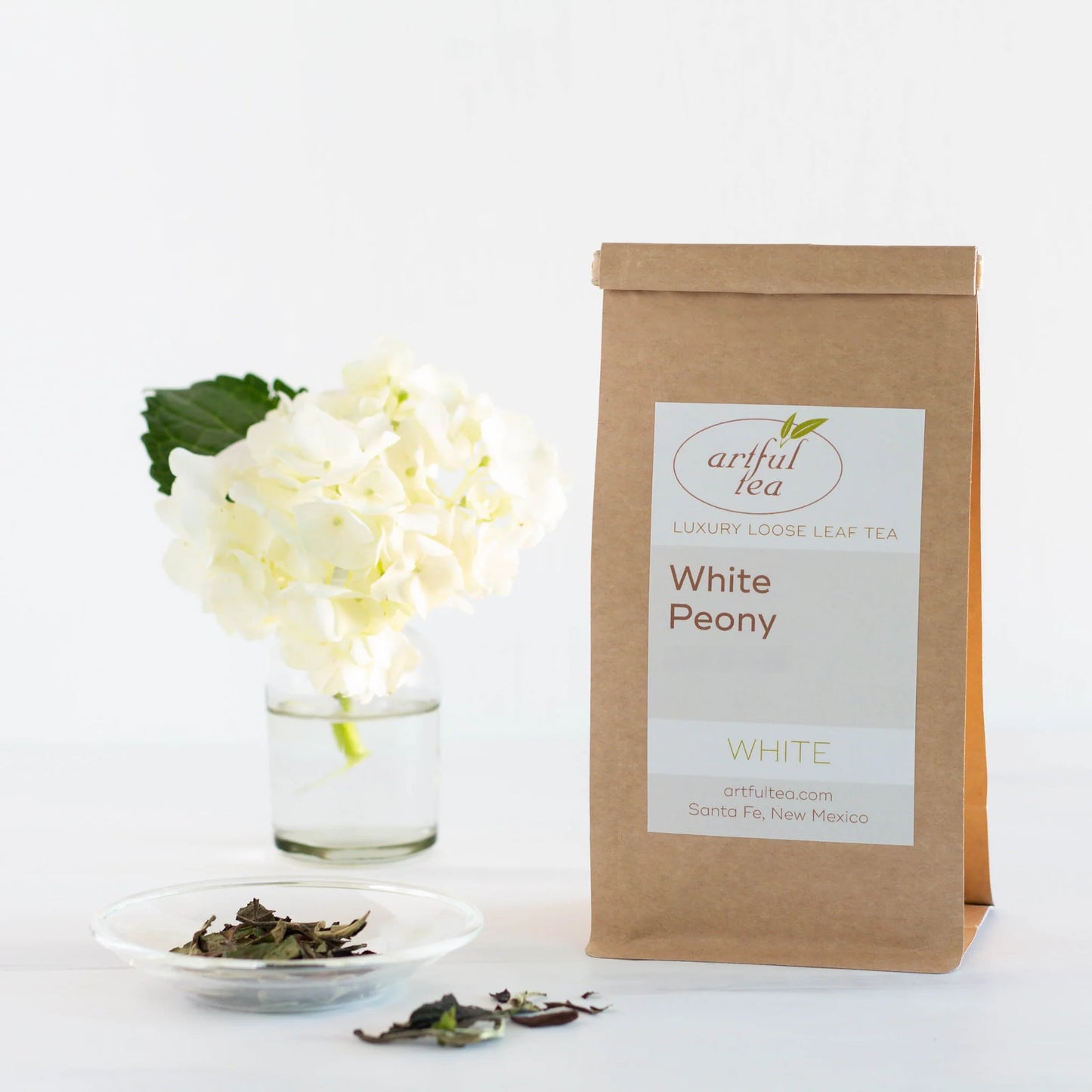 White Peony White Tea shown packaged in a brown Kraft bag with a clear dish of loose tea leaves next to it as well as a clear vase of white flowers.