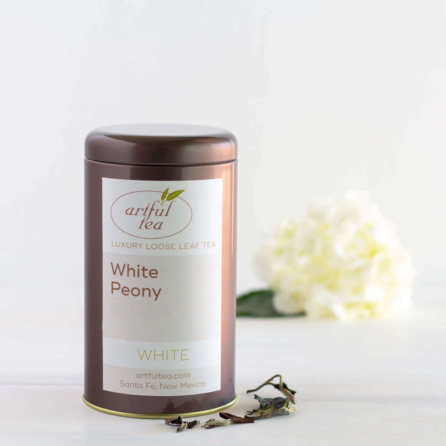 White Peony White Tea shown packaged in a brown tin with loose tea leaves next to it. In the background is a bundle of white flowers.