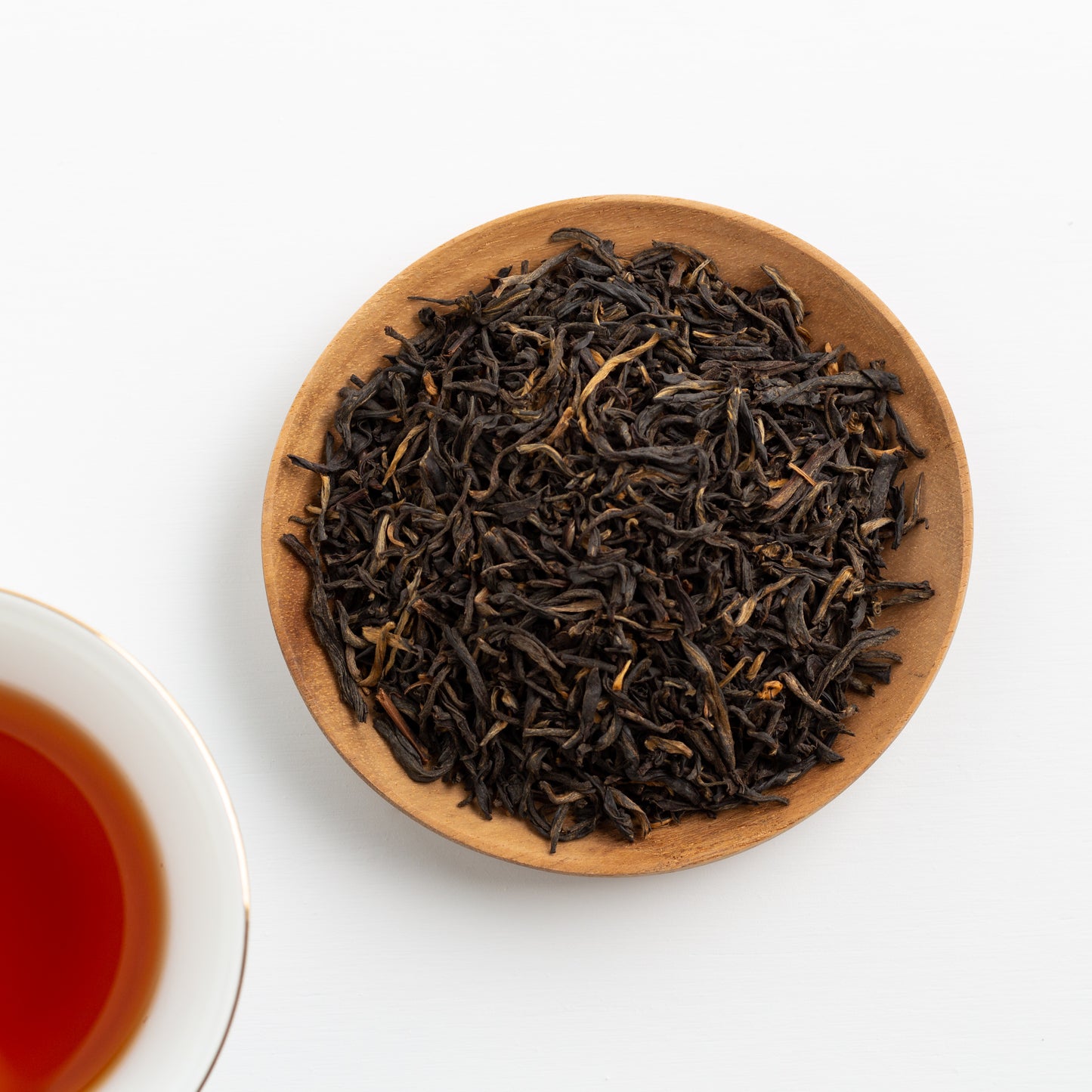 Golden Yunnan Organic Black Tea shown from above a loose tea leaves in a small wooden dish. Brewed tea in a white cup is in the lower left corner