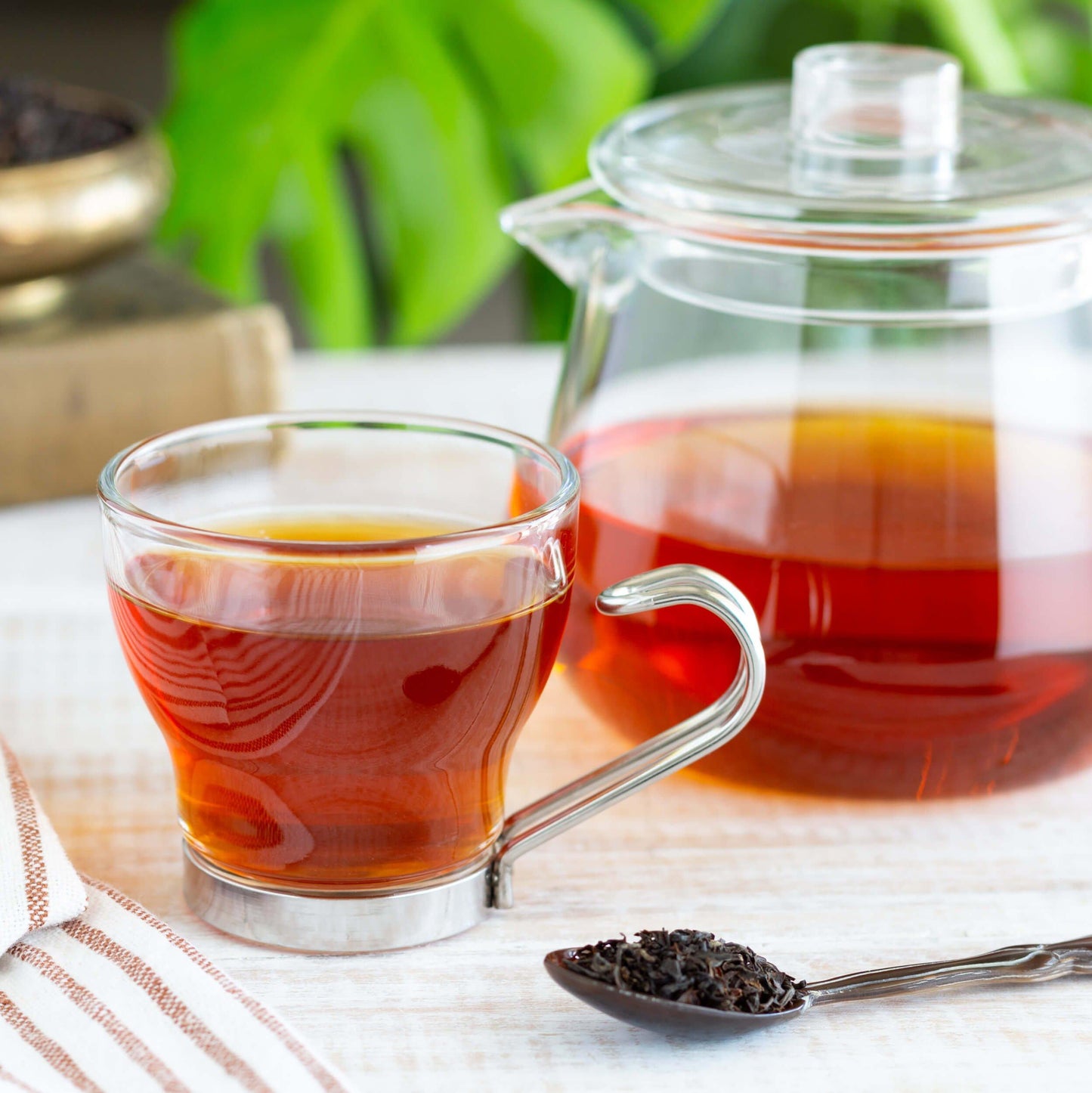 Assam Organic Black Tea shown brewed in a glass mug and glass teapot next to a spoon full of loose tea leaves.