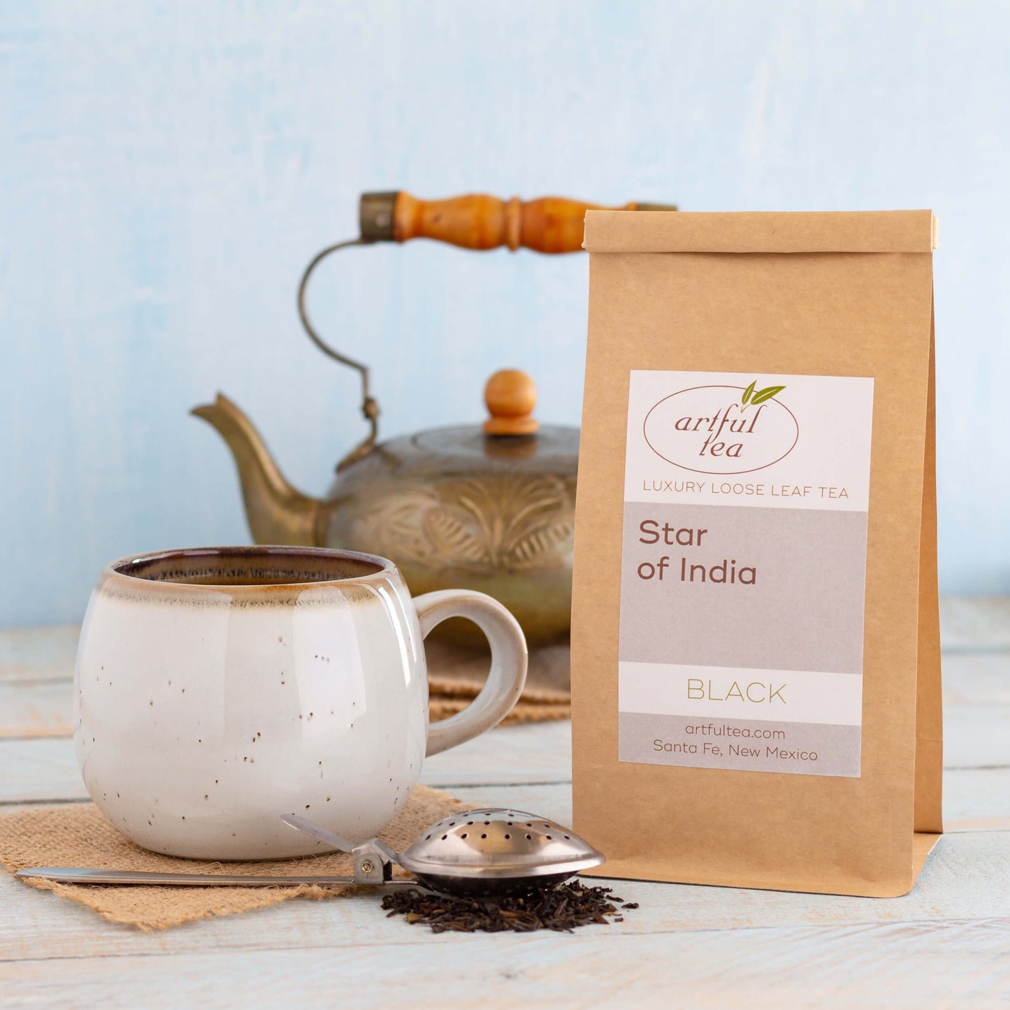 Star of India Black Tea shown packaged in a brown Kraft bag next to a white speckled mug and silver tea strainer full of loose tea leaves. In the background is a golden teapot.