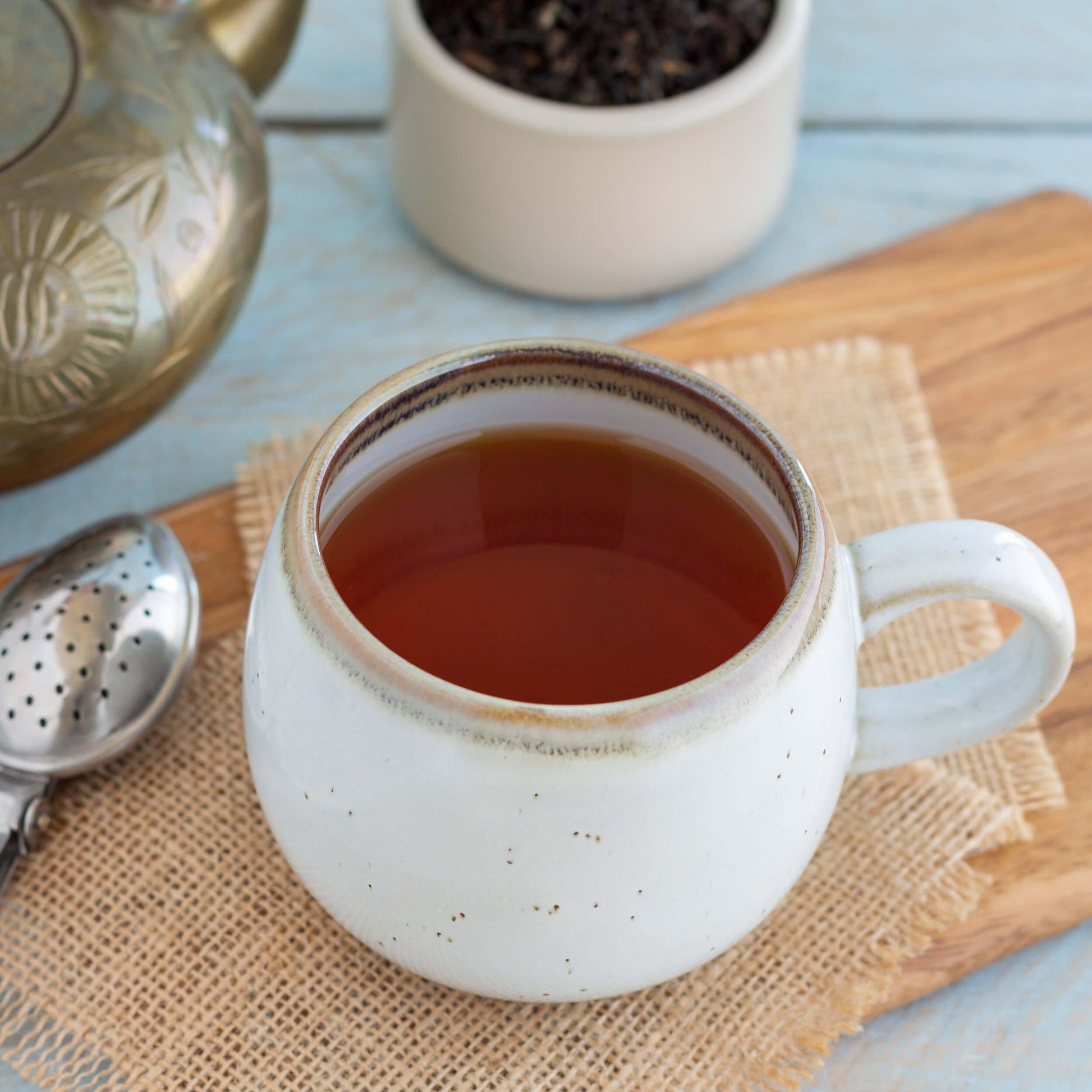 Star of India Black Tea shown as brewed tea in a round white  speckled mug, displayed on squares of burlap on top of a wooden board, with a metal tea infuser nearby