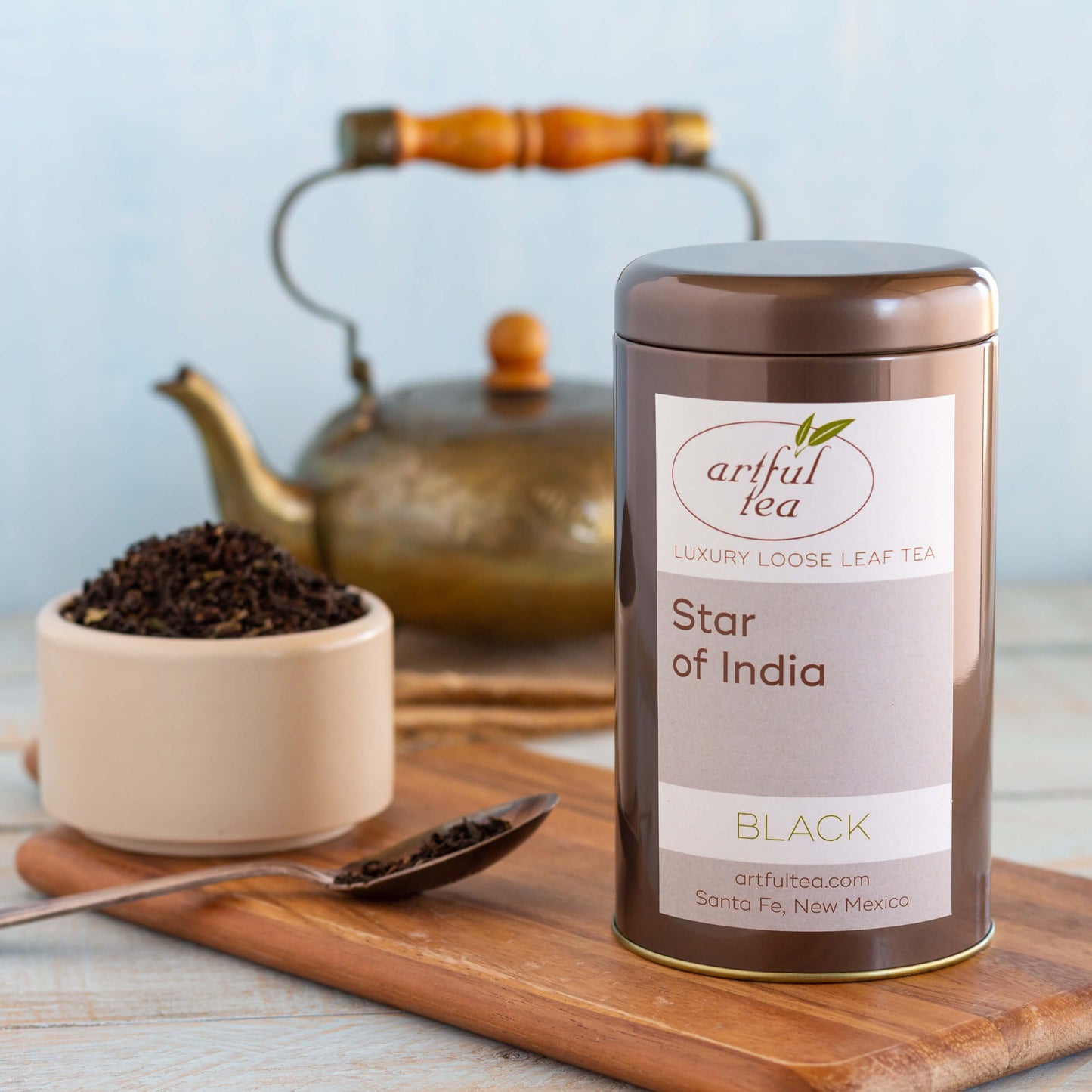 Star of India Black Tea shown packaged in a brown tin on a wooden plank. Next to it is a white cup and spoon full of loose tea leaves and in the background is a golden tea pot.