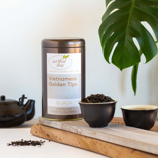 Vietnamese Golden Tips Black Tea is shown in a brown tin on top of a wooden plank. Next to it are two black cups with white interior, one full of loose tea leaves the other full of brewed tea, as well as a black tea pot in the background.
