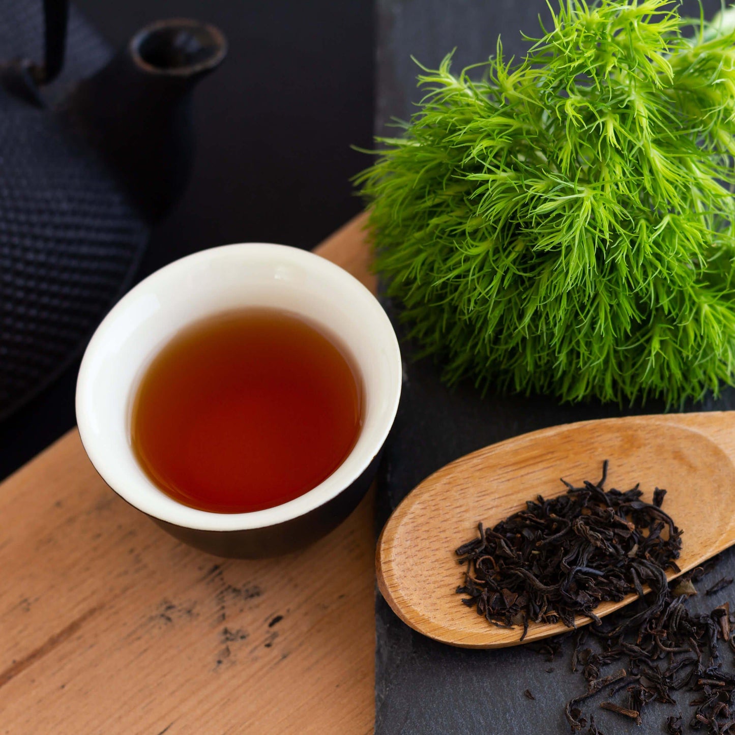 Japanese Wakoucha Organic Black Tea shown as brewed tea in a small black teacup with white interior. A leafy green plant is nearby, as well as a wooden spoon with loose tea leaves. The background is dark.