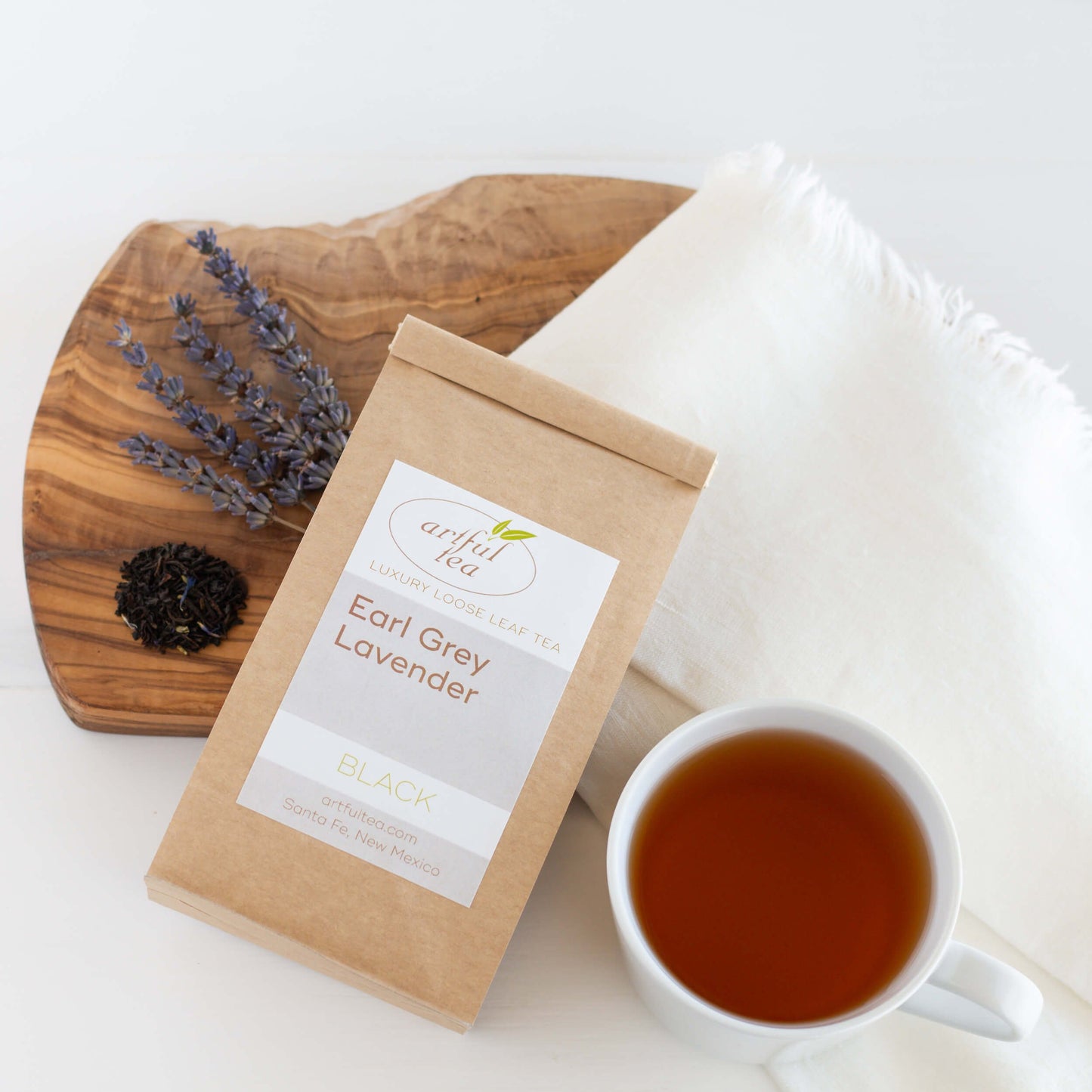 Earl Grey Lavender Black Tea shown packaged in a brown Kraft bag on a white cloth and wooden plank. A white mug full of brewed tea and ingredients nearby.