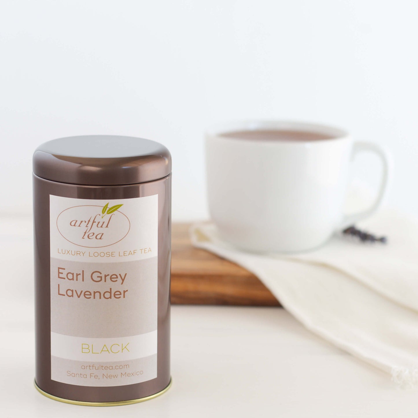 Earl Grey Lavender Black Tea shown packaged in a brown tin. In the background is a white mug full of brewed tea on a white cloth and wooden plank.