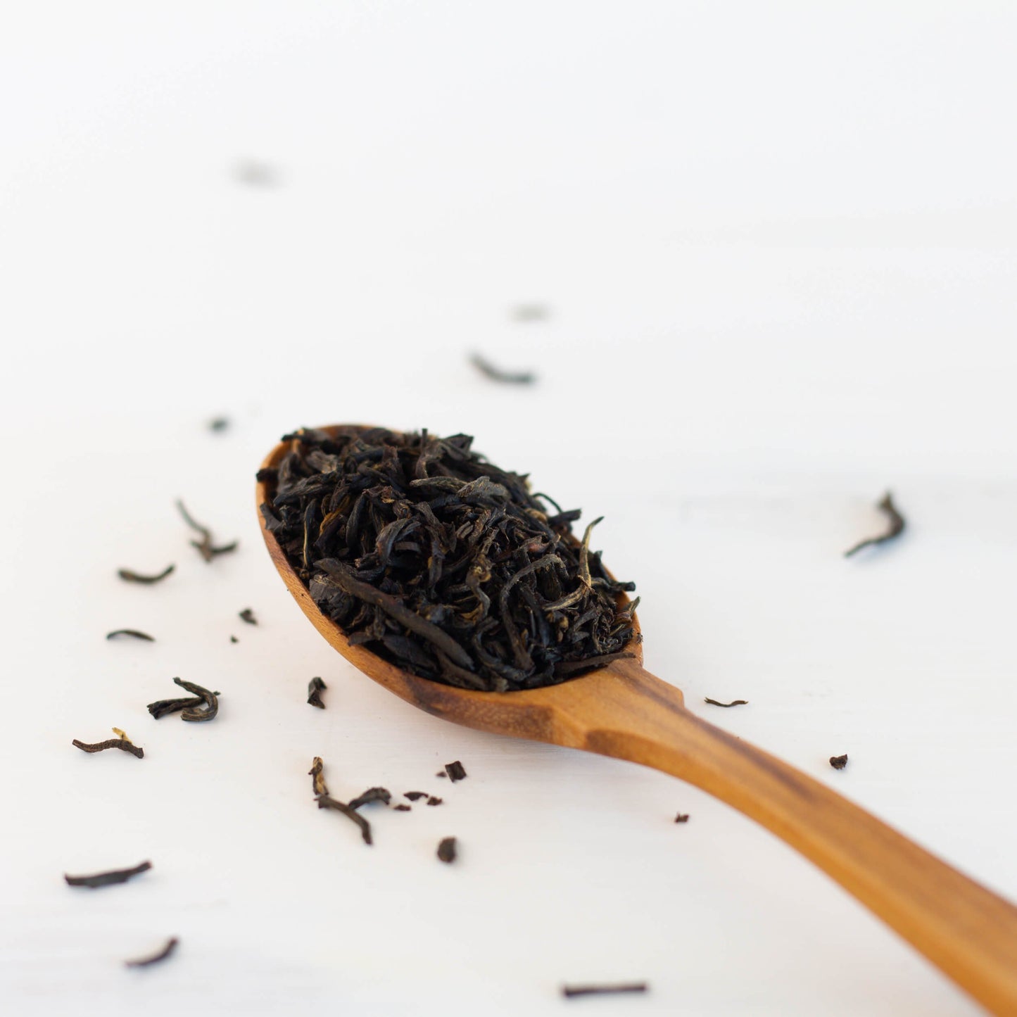 Earl Grey loose black tea leaves shown on a wooden spoon with loose tea leaves dispersed around it on a white background.