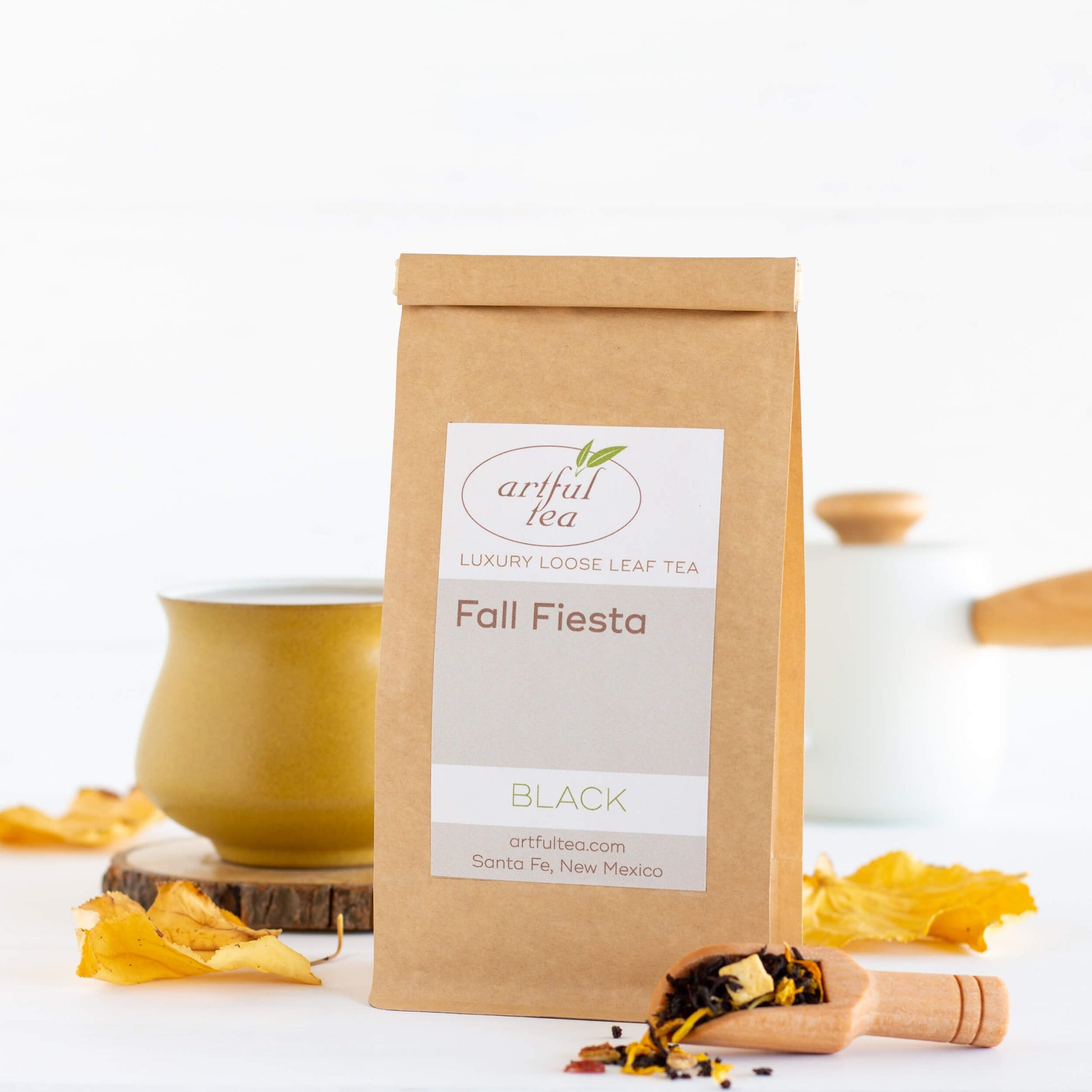 Fall Fiesta Black Tea shown packaged in a brown Kraft bag alongside a mustard yellow mug on top of a wooden coaster, and a scoop of loose tea leaves.