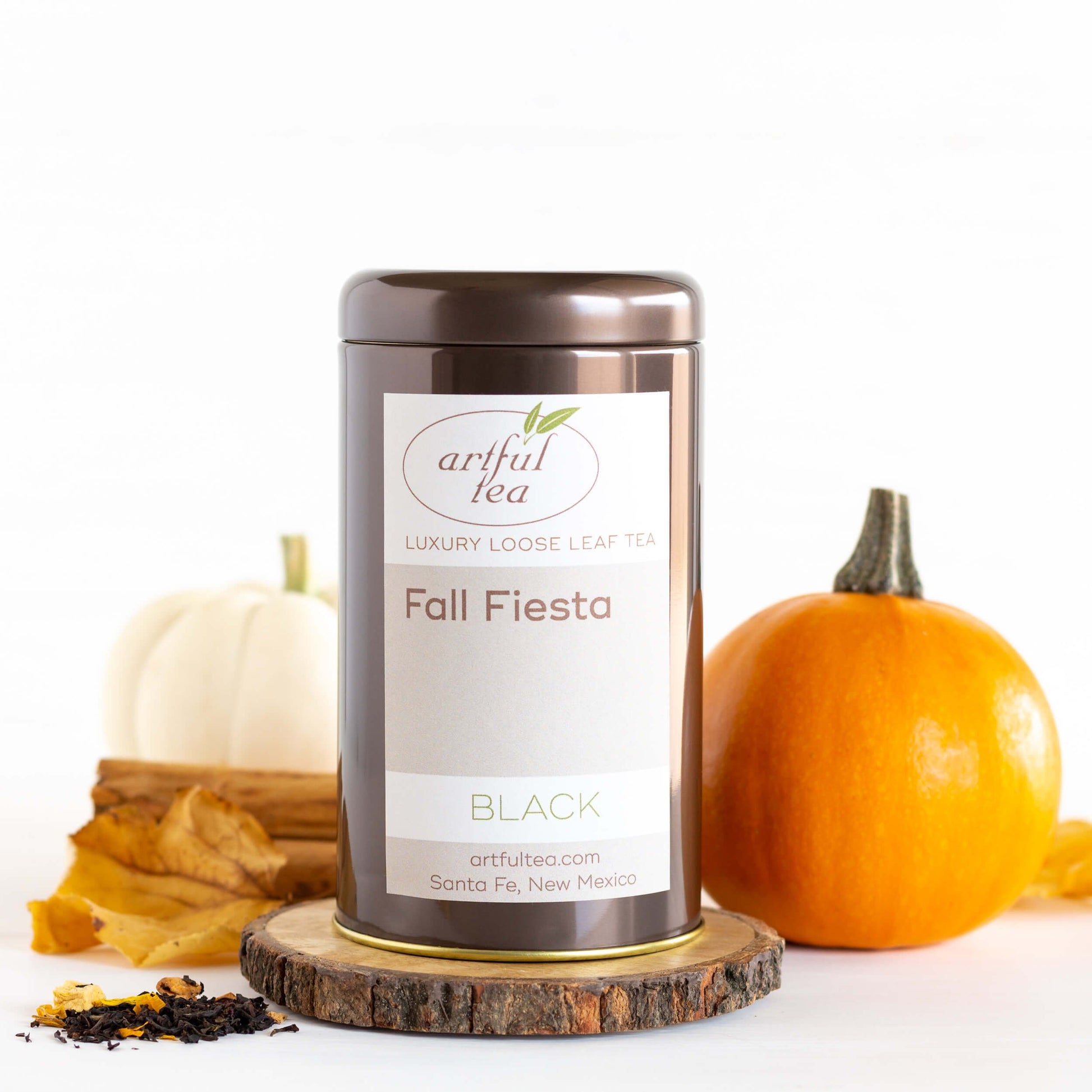 Fall Fiesta Black Tea shown packaged in a brown tin on top of a wooden coaster. Loose tea leaves and pumpkins are nearby.