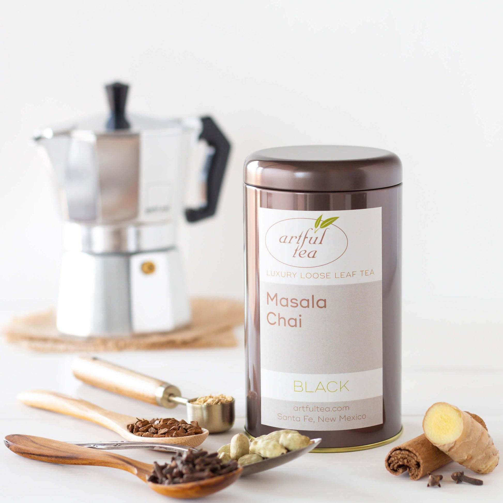 Masala Chai Black Tea shown packaged in a brown tin with four spoons full of spices nearby, and a chrome moka pot in the background