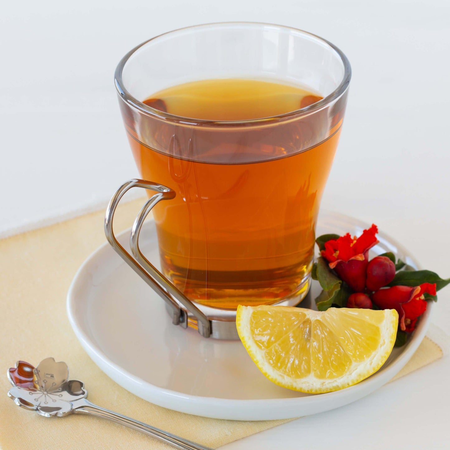 Pomegranate Lemon Black Tea shown as brewed tea in a glass mug with a metal handle, on a white dish with a lemon wedge and red blossoms.