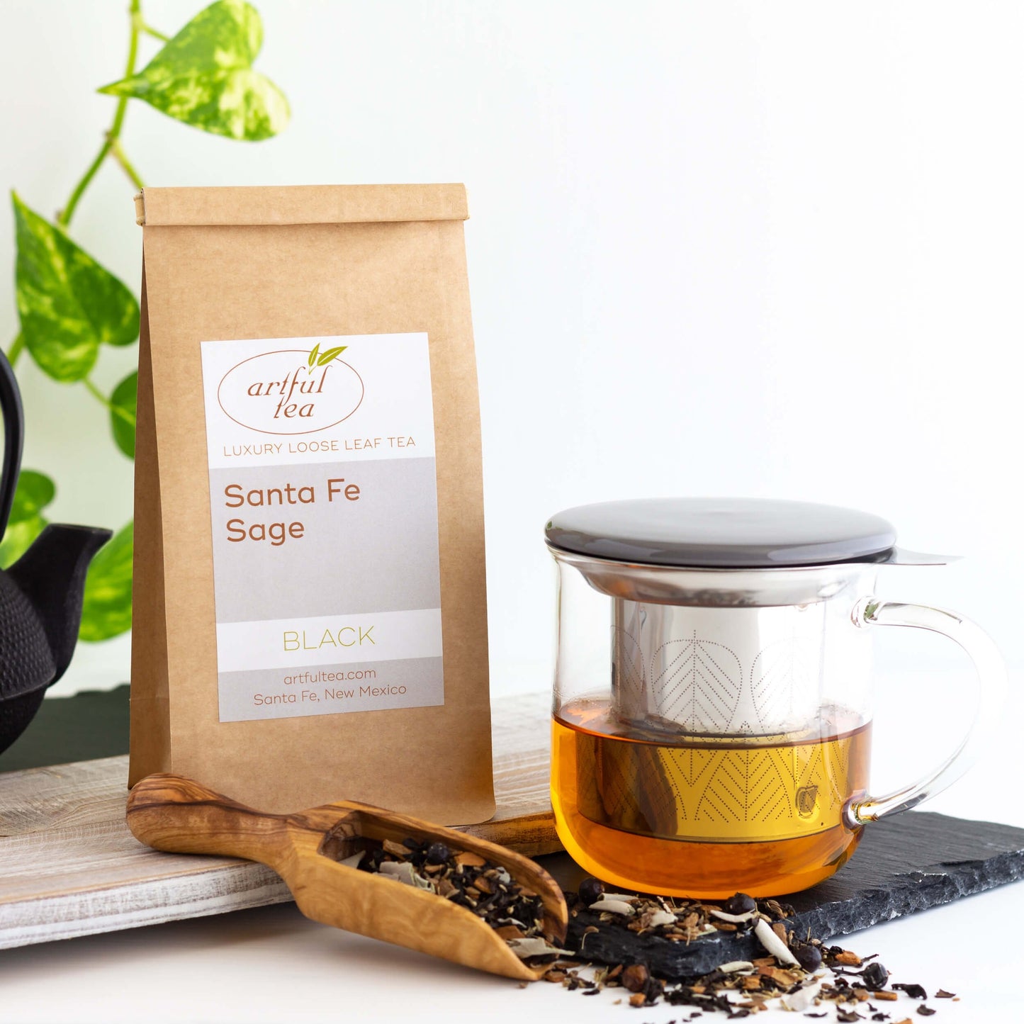 Santa Fe Sage black tea brewed in a clear mug alongside a scoop with loose tea leaves scattered around. Next to it is a brown Kraft bag of Santa Fe Sage black tea on a wooden plank.