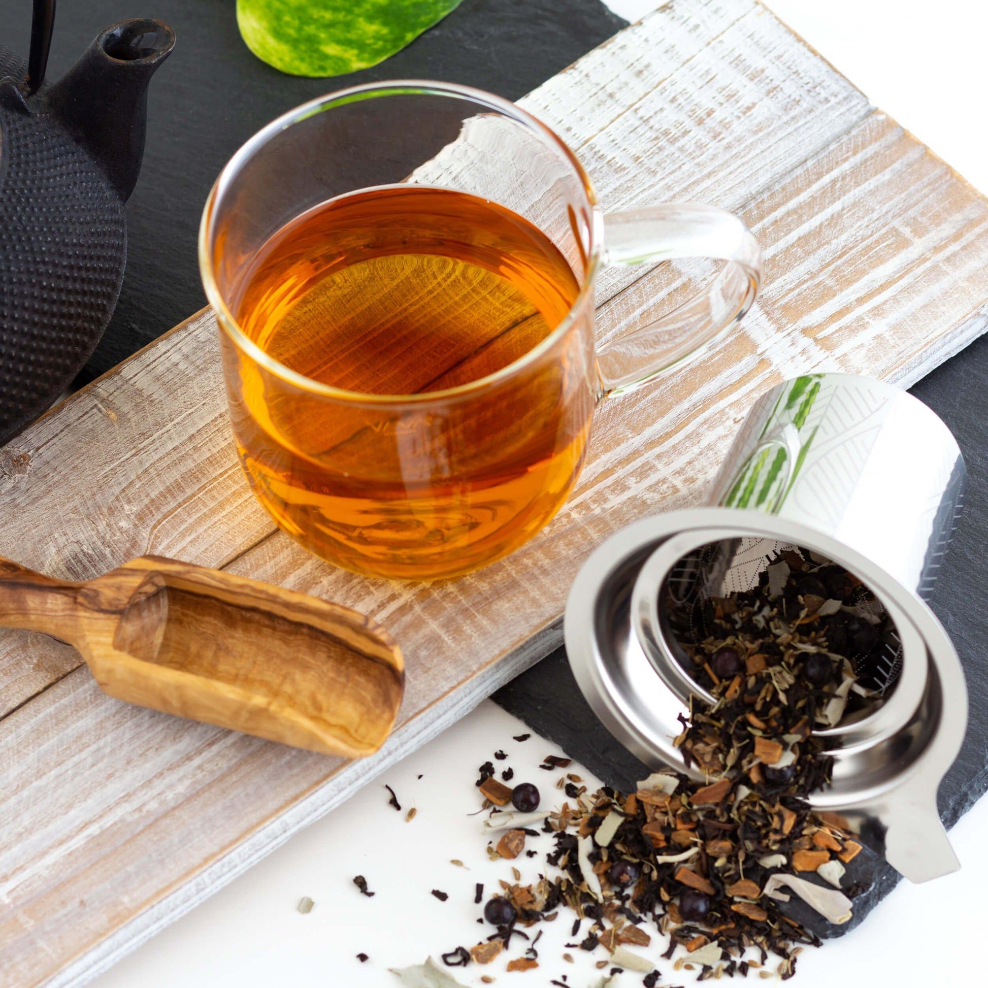 Santa Fe Sage Black Tea shown as brewed tea in a glass mug on a whitewashed wooden plank. A metal infuser is on its side with loose leaf tea spilling out, and a wooden scoop is nearby.