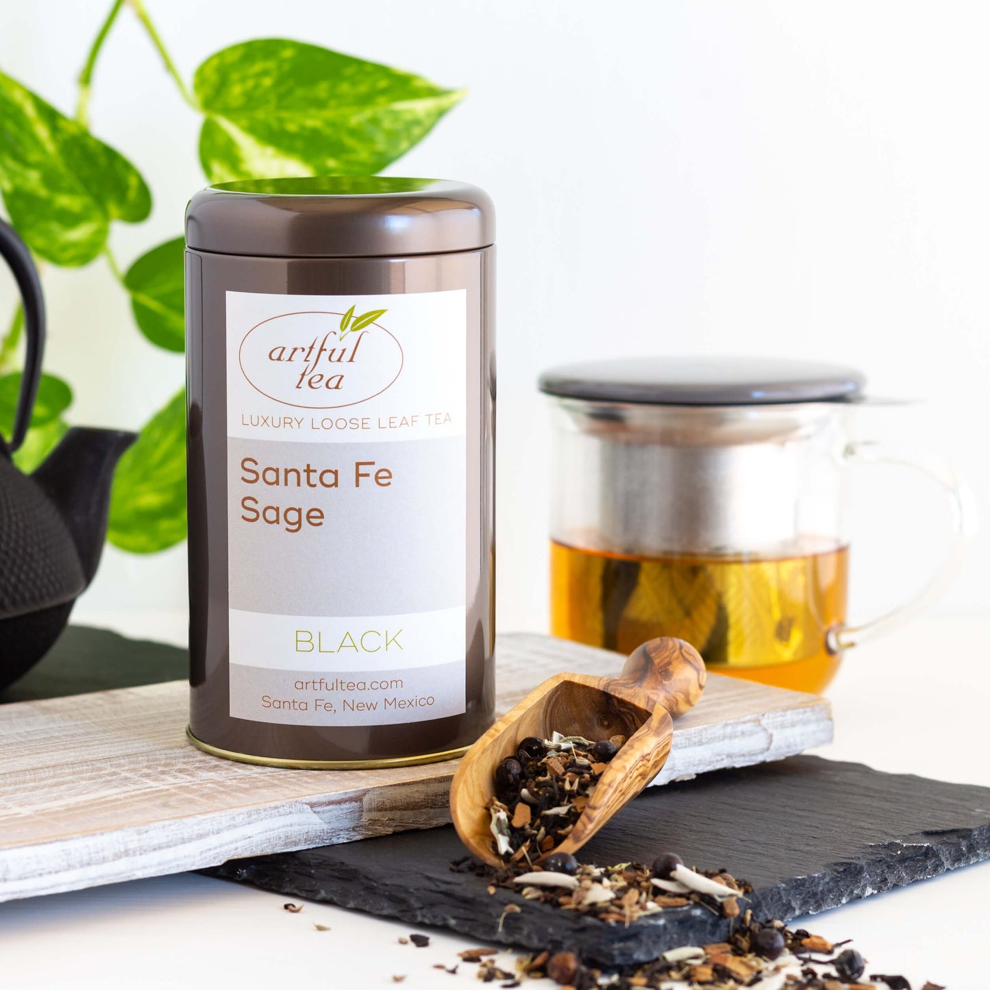 Santa Fe Sage black tea in a brown tin alongside a scoop with loose tea leaves scattered around, as well as a mug full of brewed tea in the background.