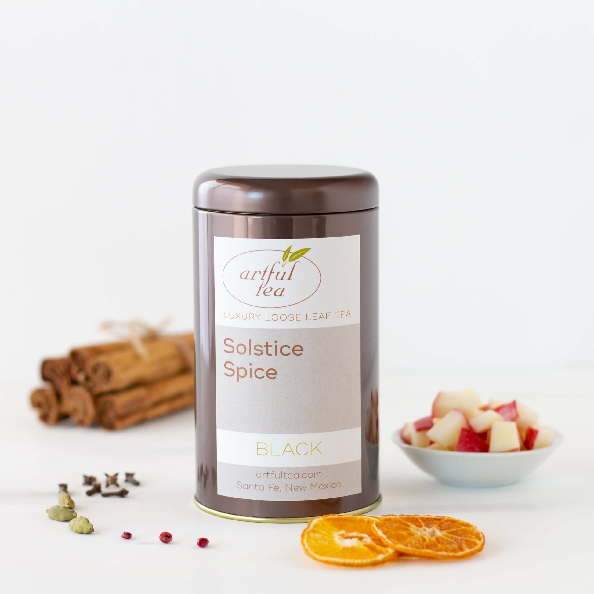 Solstice Spice Black Tea shown packaged in a brown tin with ingredients such as dried oranges, spices, apples, and cinnamon sticks surrounding it.