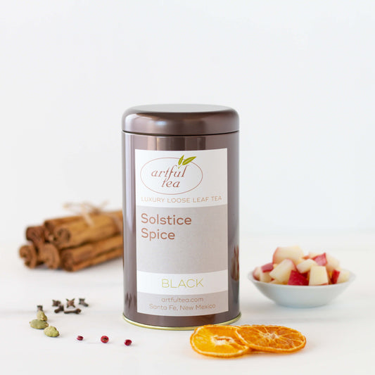 Solstice Spice Black Tea shown packaged in a brown tin with ingredients such as dried oranges, spices, apples, and cinnamon sticks surrounding it.