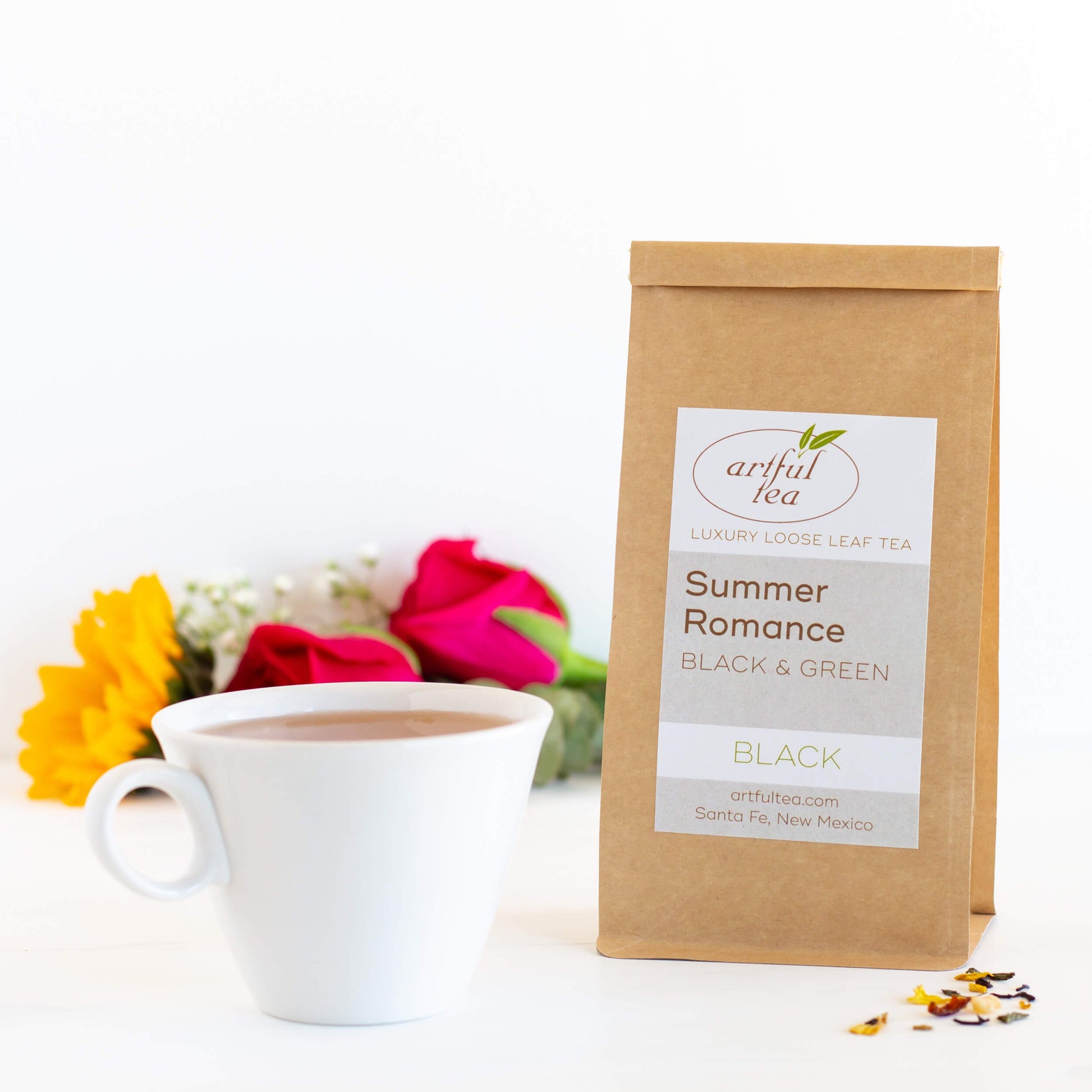 Summer Romance Black and Green Tea blend shown packaged in a brown Kraft bag next to a white mug full of brewed tea. In the background is a bundle of white, red, and yellow flowers.