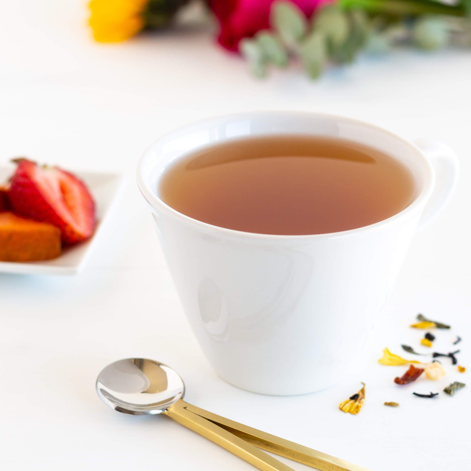 Summer Romance Black & Green Tea shown as brewed tea in a small white cup with strawberries and flowers in the background, and a brass handled spoon in the foreground.