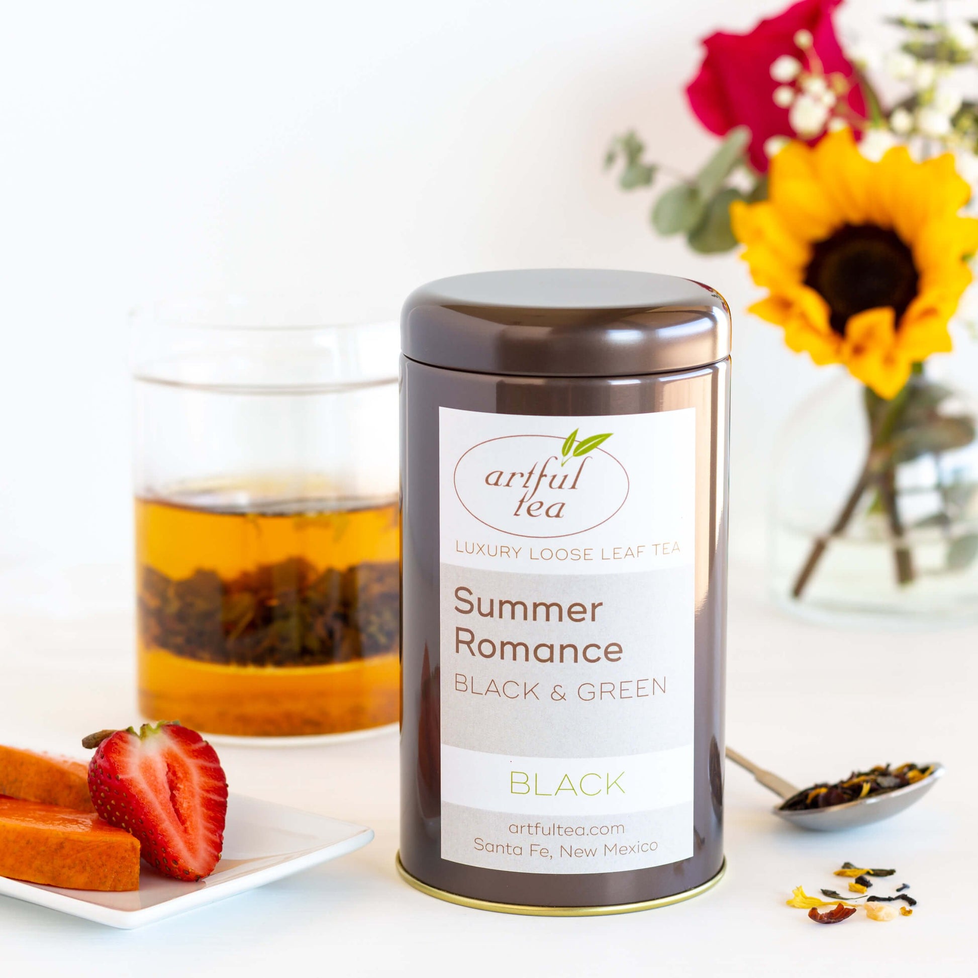 Summer Romance Black and Green Tea Blend shown packaged in a brown tin. Next to it is a plate of fruit, a spoon of loose tea leaves, and a clear cup of brewed tea and steeped leaves. In the background is a vase of red and yellow flowers.