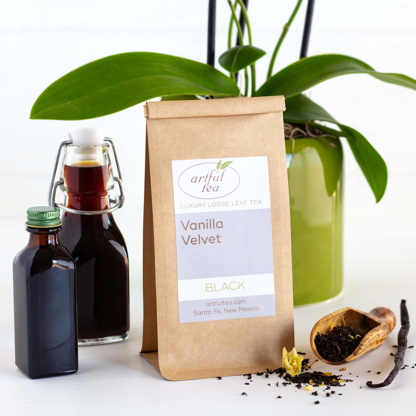 Vanilla Velvet Black Tea shown packaged in a brown Kraft bag. Next to it are two clear bottles of vanilla syrup and a wooden scoop with loose tea leaves spilling out, along with a stick of vanilla.