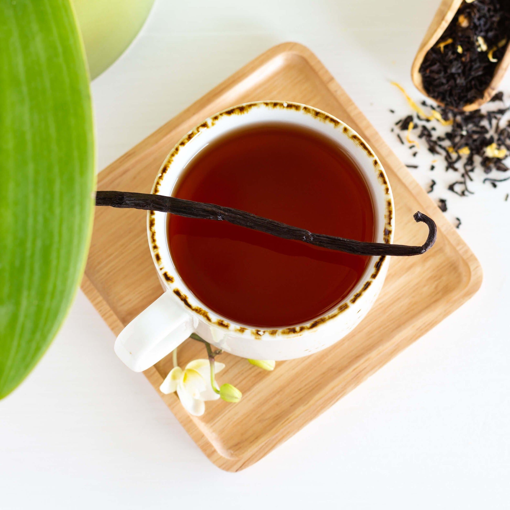 Vanilla Velvet Black Tea shown from above as brewed tea in a white mug with brown rim, displayed on a square bamboo coaster, with a vanilla bean lying across the top of the mug.
