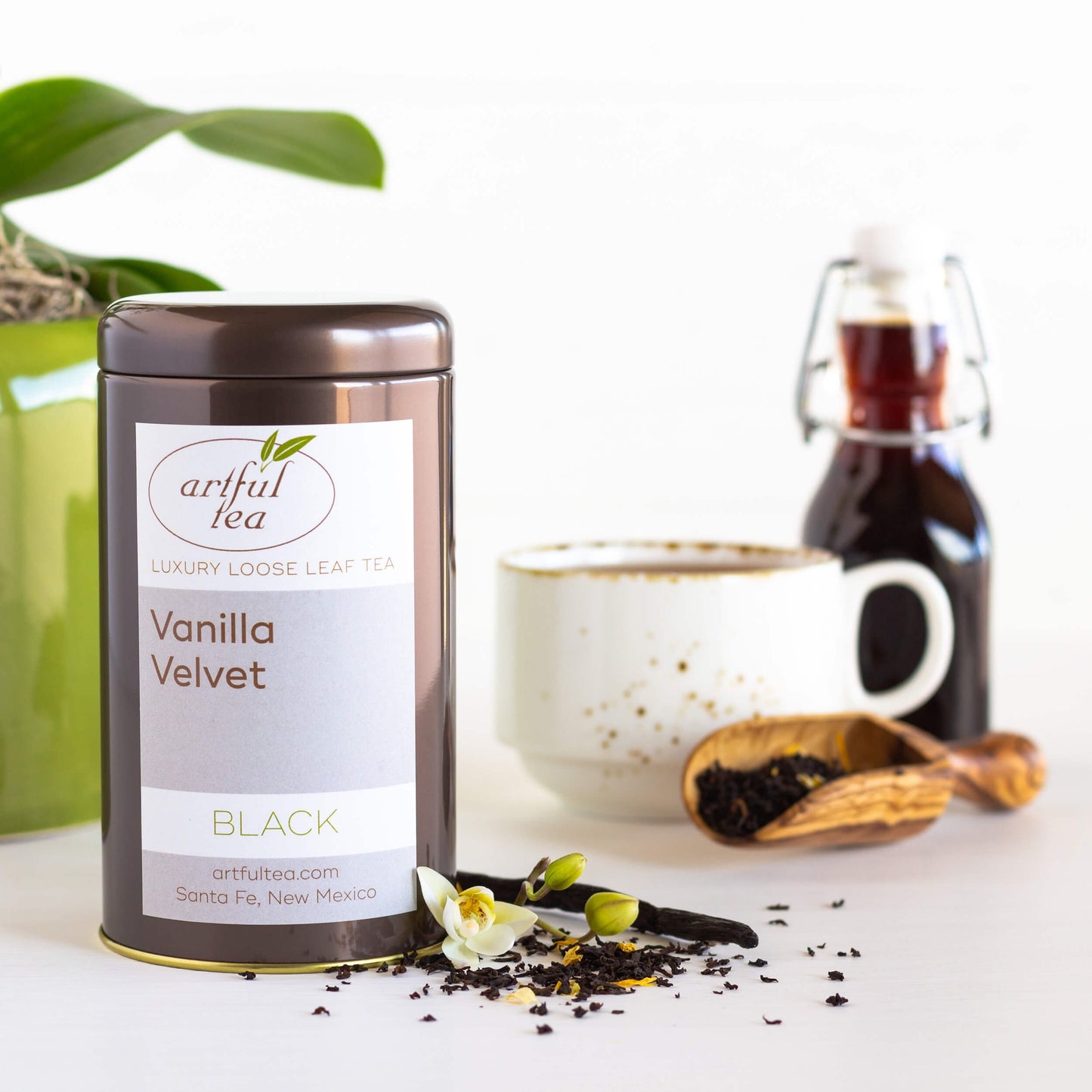 Vanilla Velvet Black Tea shown packaged in a brown tin with loose tea leaves and a vanilla stick strewn next to it. In the background is a white mug full of brewed tea, a wooden scoop full of loose tea leaves, and a clear bottle full of vanilla syrup.