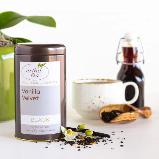 Vanilla Velvet Black Tea shown packaged in a brown tin with loose tea leaves and a vanilla stick strewn next to it. In the background is a white mug full of brewed tea, a wooden scoop full of loose tea leaves, and a clear bottle full of vanilla syrup.