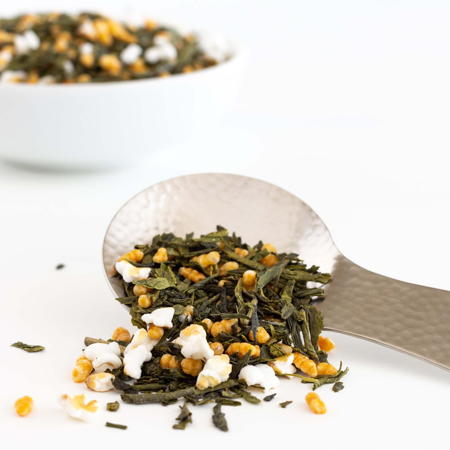 Genmaicha Organic Green Tea. Loose leaves, including roasted brown rice kernals, shown overflowing a stainless steel tea scoop.