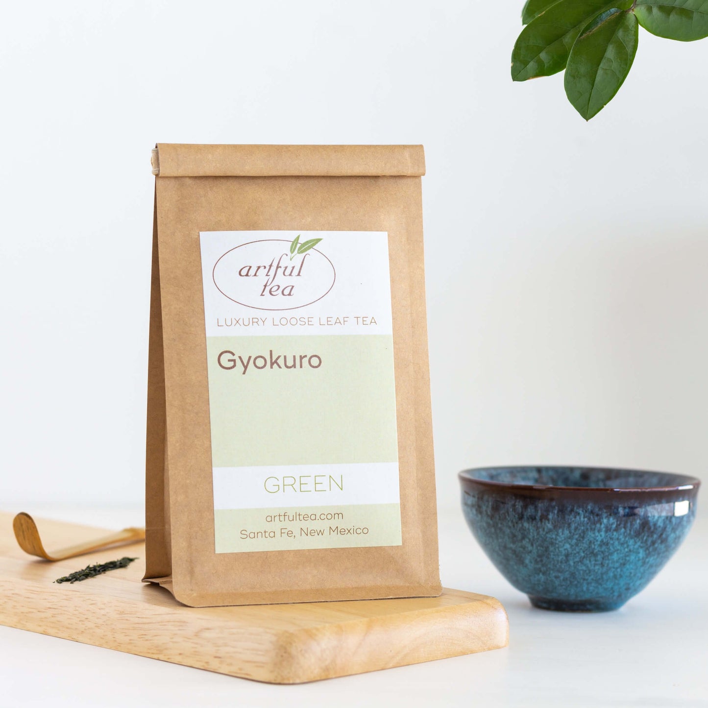 Gyokuro Green Tea shown packaged in a brown Kraft bag next to a small wooden scoop and loose tea leaves on top of a wooden plank beside a blue teacup.