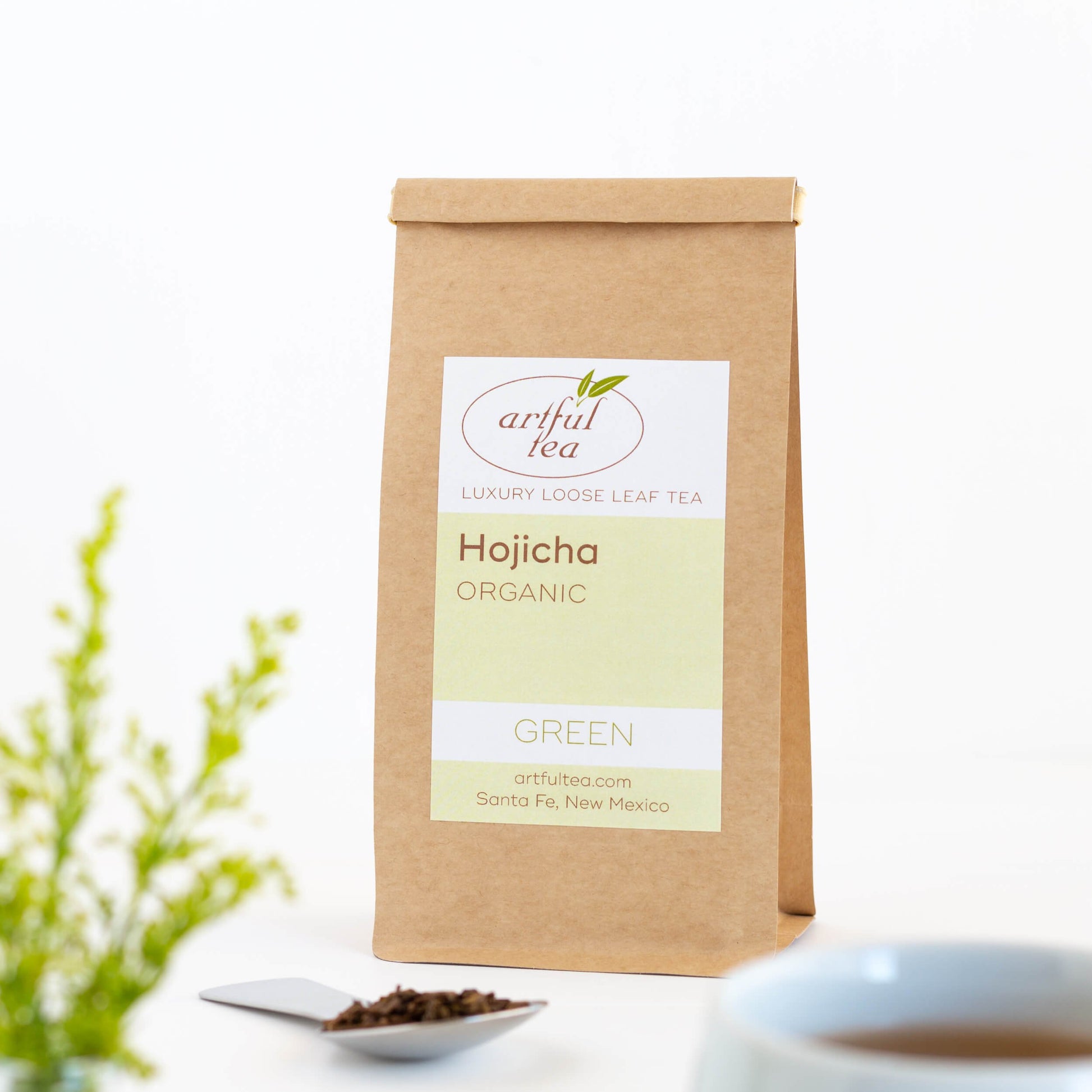 Hojicha Roasted Green Tea shown packaged in a brown Kraft bag with a silver spoon of loose leaf tea next to it.