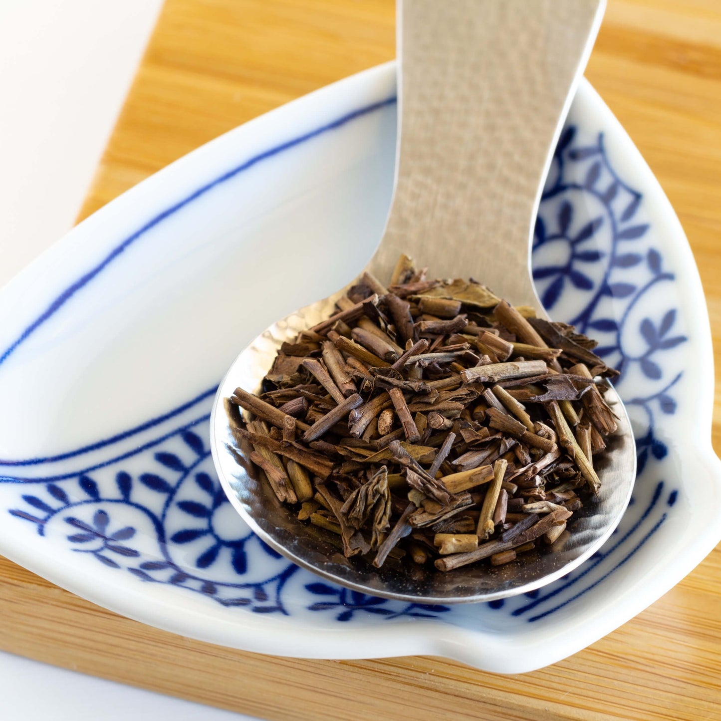 Hojicha Green Tea shown as loose leaf tea on a silver spoon on top of a white and blue dish.