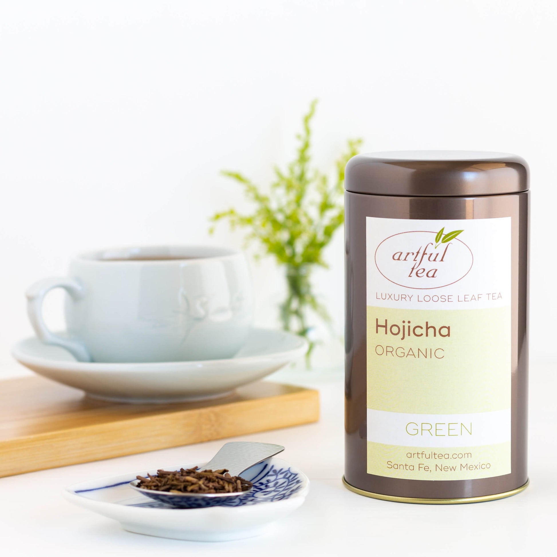 Hojicha Green Tea shown packaged in a brown tin beside a white and blue dish with a silver spoon of loose tea leaves. In the background is a white mug full of brewed tea on a white saucer, on top of a wooden plank.