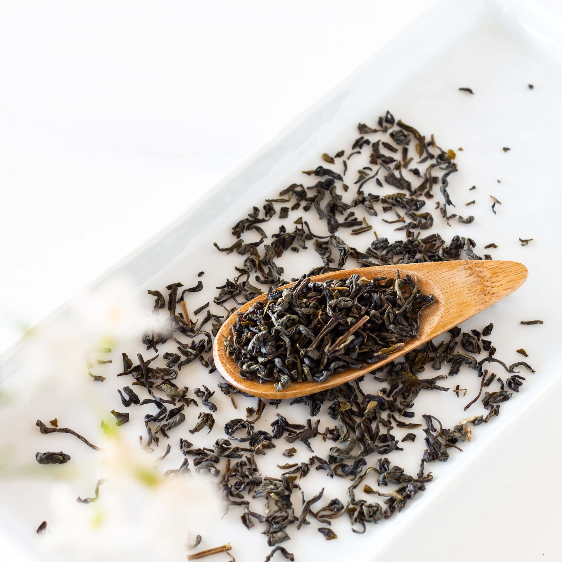 Jasmine Yin Cloud Green Tea shown as loose leaf tea inside of a wooden scoop with loose tea leaves surrounding it on a white, rectangular tray.