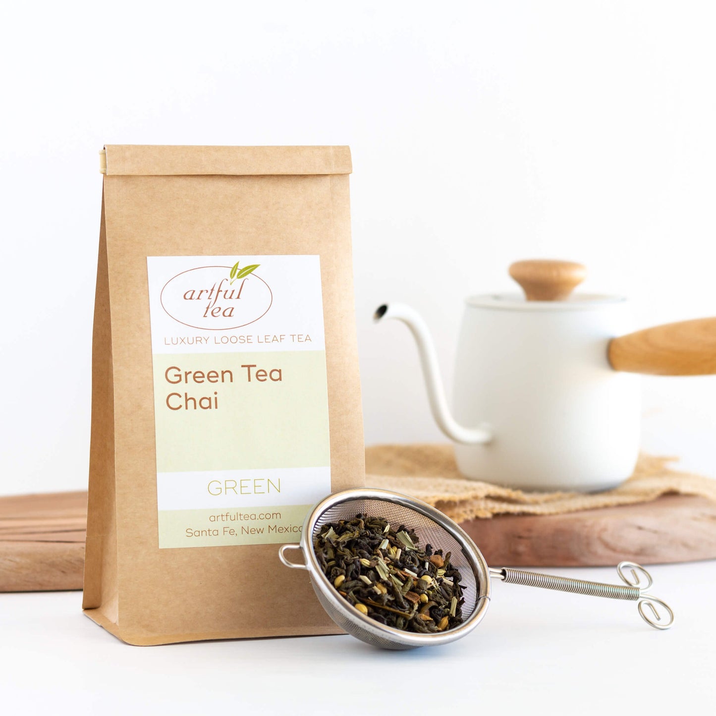 Green Tea Chai shown packaged in a brown Kraft bag next to a strainer full of loose leaf tea. In the background is a white teapot on a wooden plank.